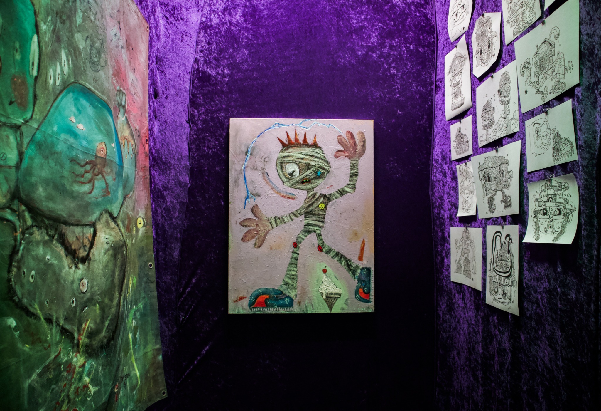Narrow art display with colorful paintings and sketches on purple walls, featuring a central abstract creature and sketches on the right.