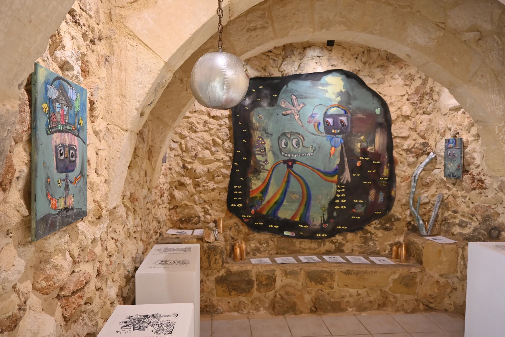 Art gallery with stone walls, featuring colorful abstract paintings and sculptures, including a large central piece and a spherical object.