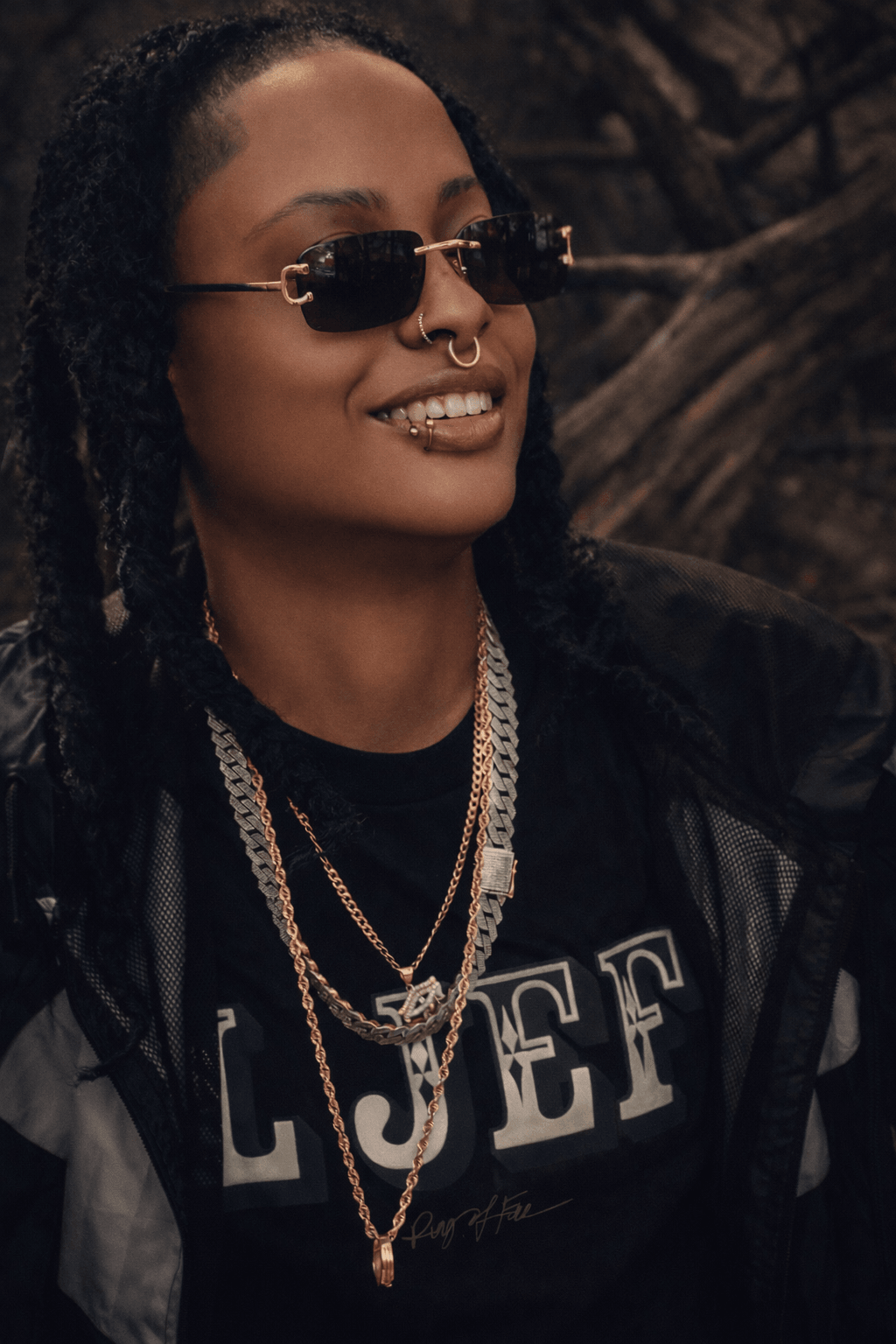Woman with sunglasses, nose piercing, layered necklaces, and a black shirt with white text, outdoors with trees in background.