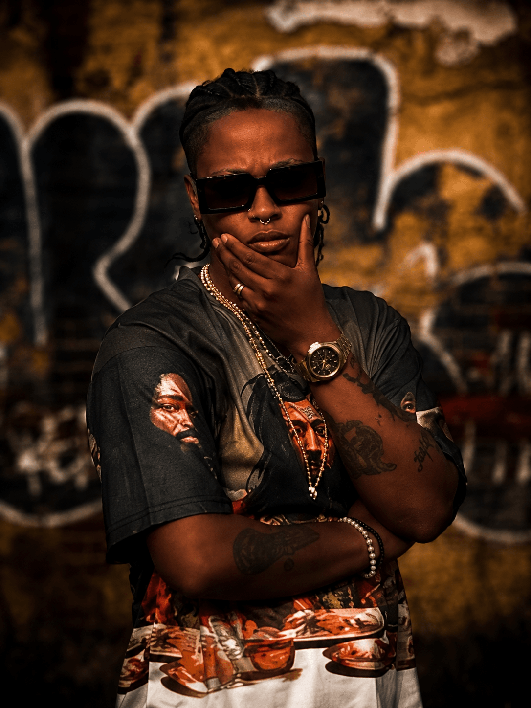 Person with sunglasses and jewelry poses with hand on chin in front of graffiti wall.