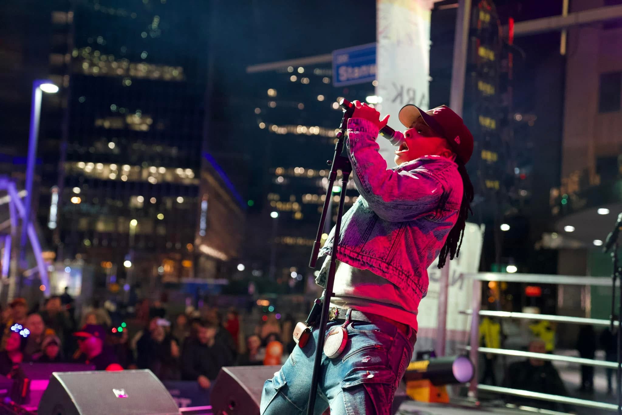 Person in pink jacket and red hat holding a microphone on stage at night with city lights and crowd in background.