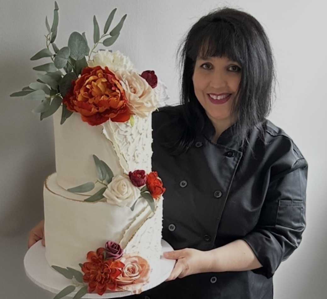 Woman with black hair and black jacket holding a large decorated cake with flowers.