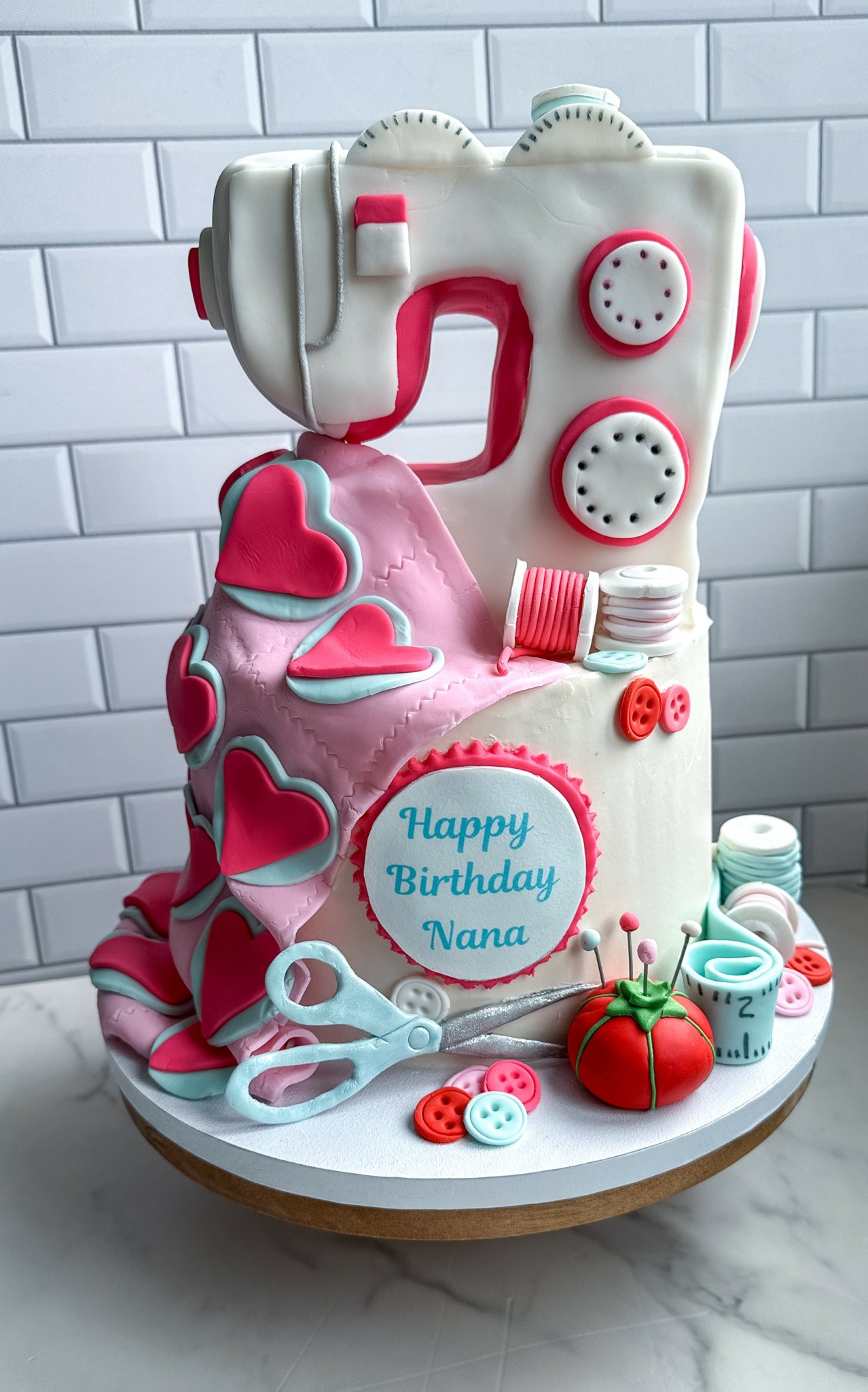 Cake shaped like a sewing machine with pink and white decorations, a blue message reading 'Happy Birthday Nana,' and sewing-themed embellishments.