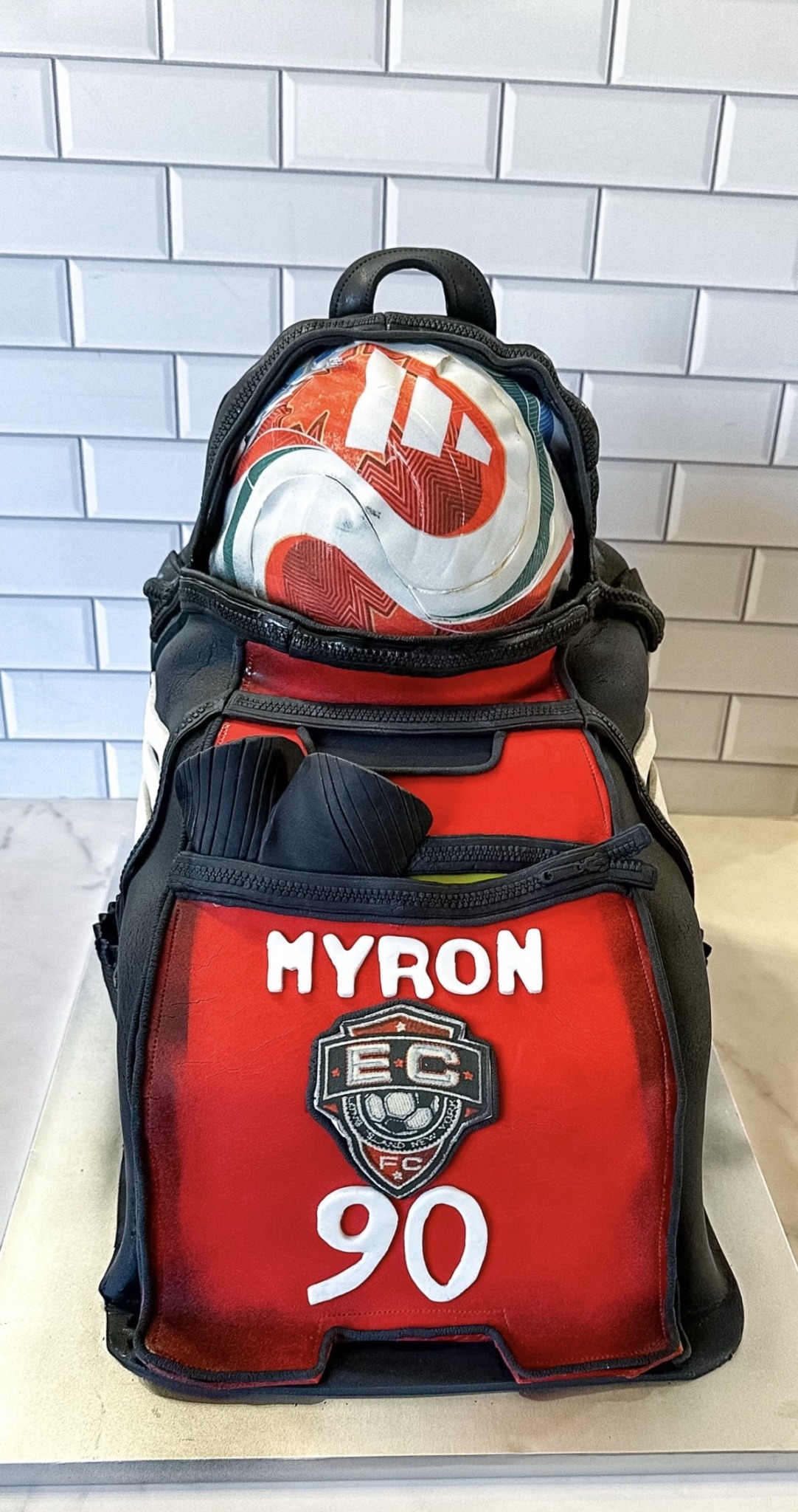 Backpack with a Nike ball on top, red and black colors, labeled MYRON, number 90, and a logo, against a tiled wall.