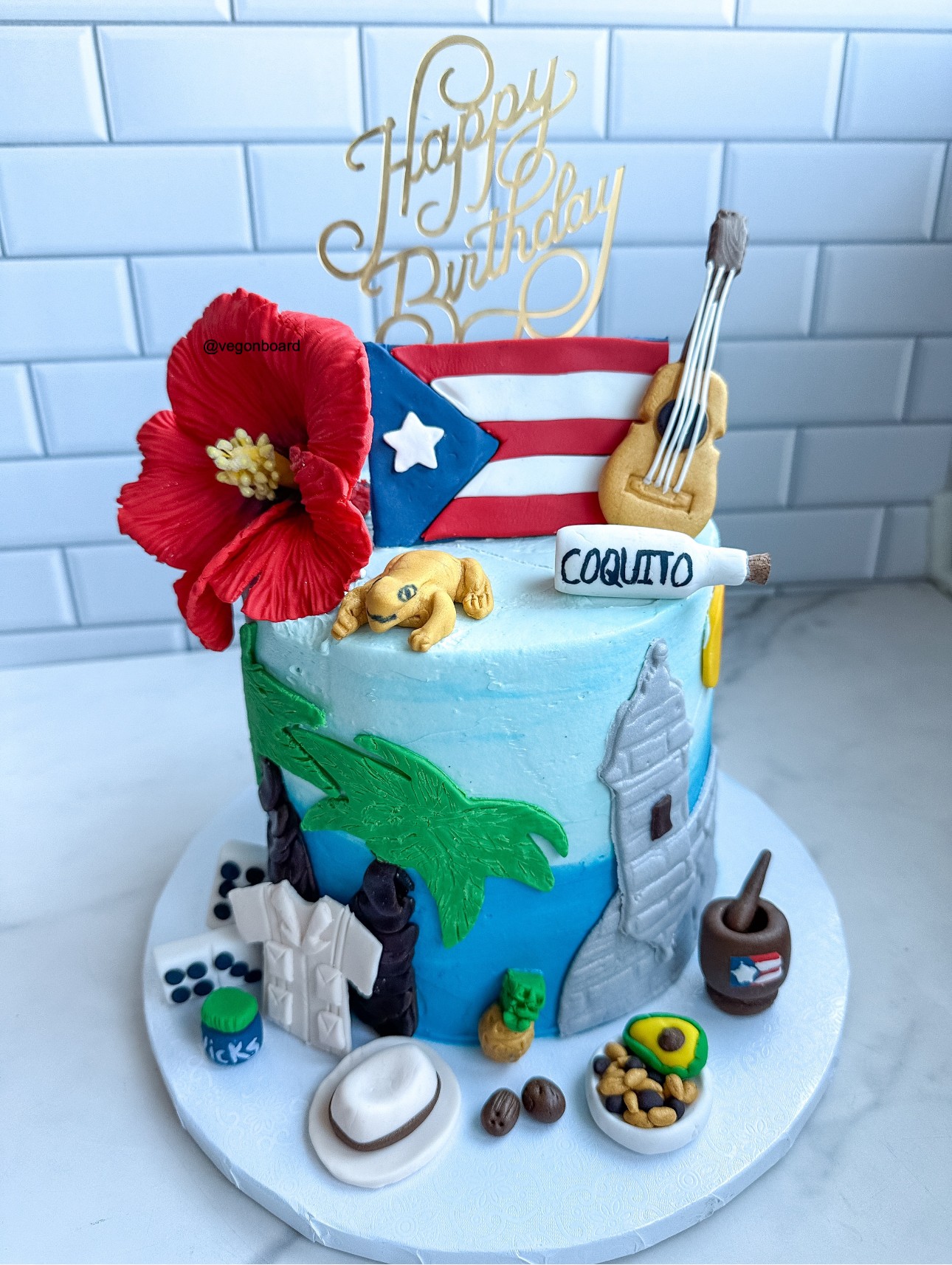 Celebration cake with a red flower, American flag, guitar, and various decorations, set against a white tiled background.