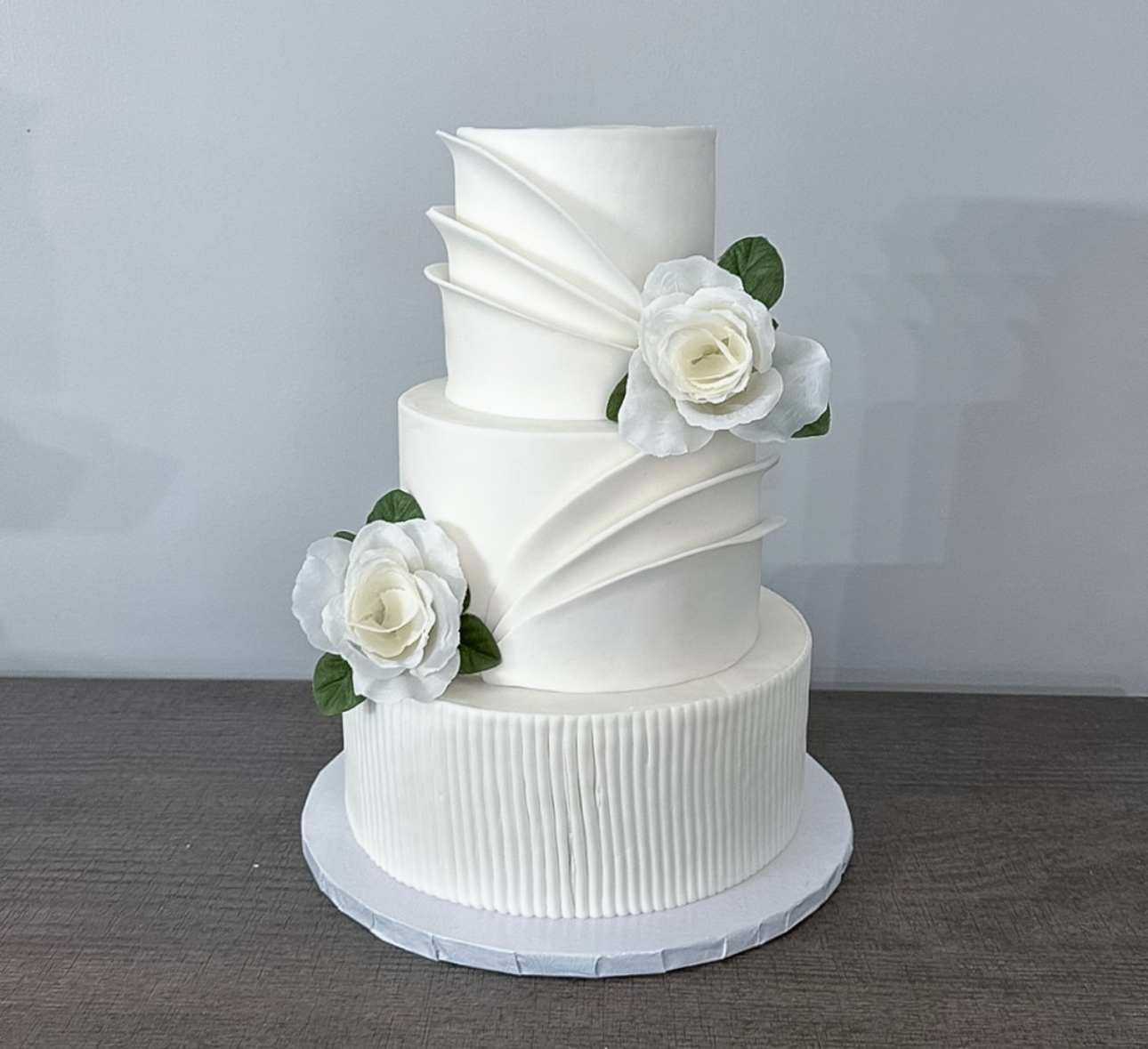 Three-tiered white wedding cake with floral decorations, placed on a gray surface against a light background.