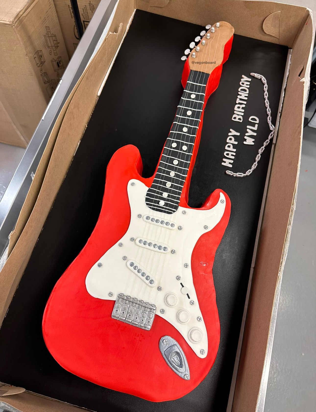 Red and white electric guitar-shaped birthday cake with a black background, decorated with a message and chain, for a birthday celebration.