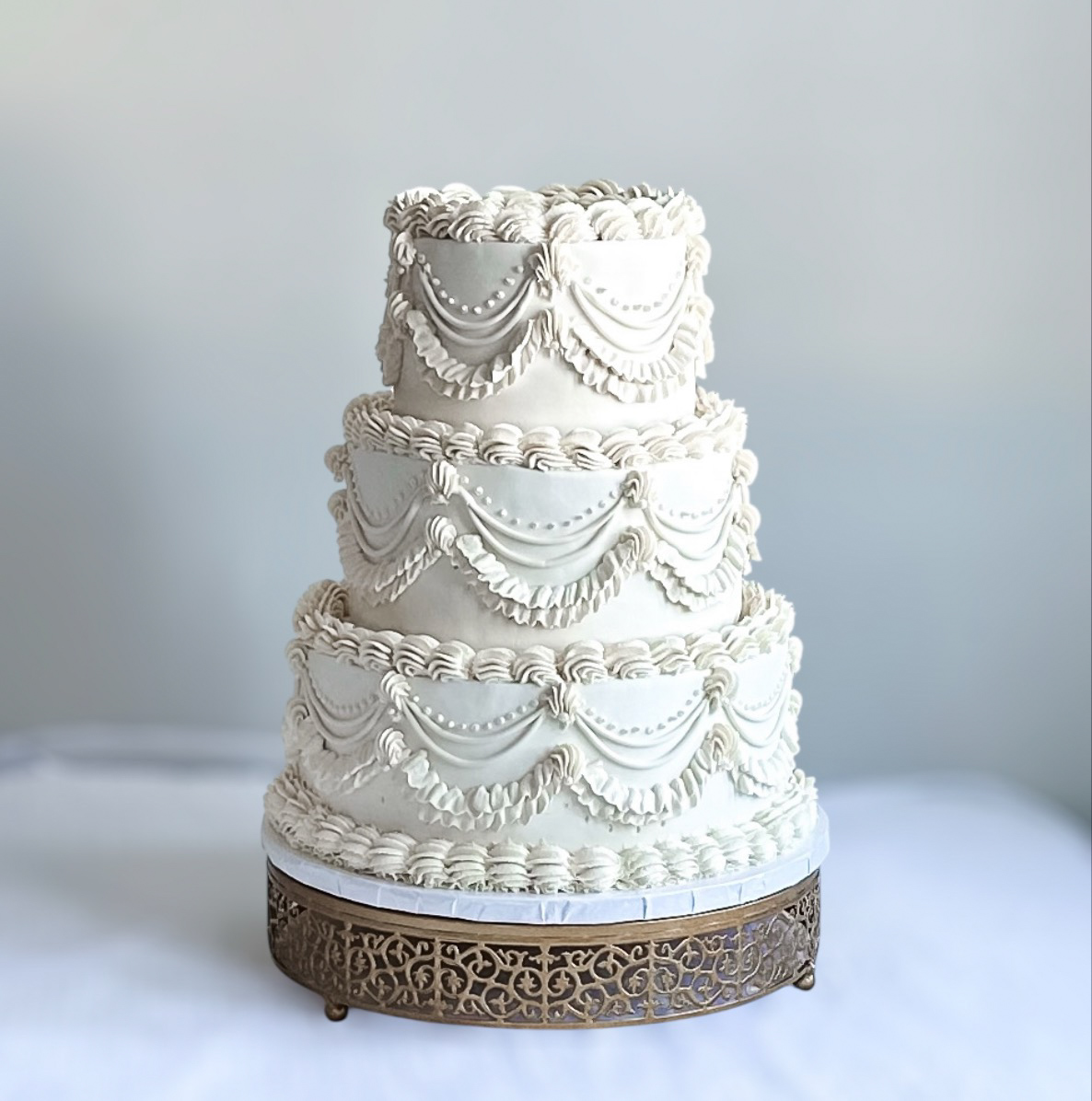 Three-tiered white wedding cake with decorative icing, on a decorative metal stand, against a plain background.