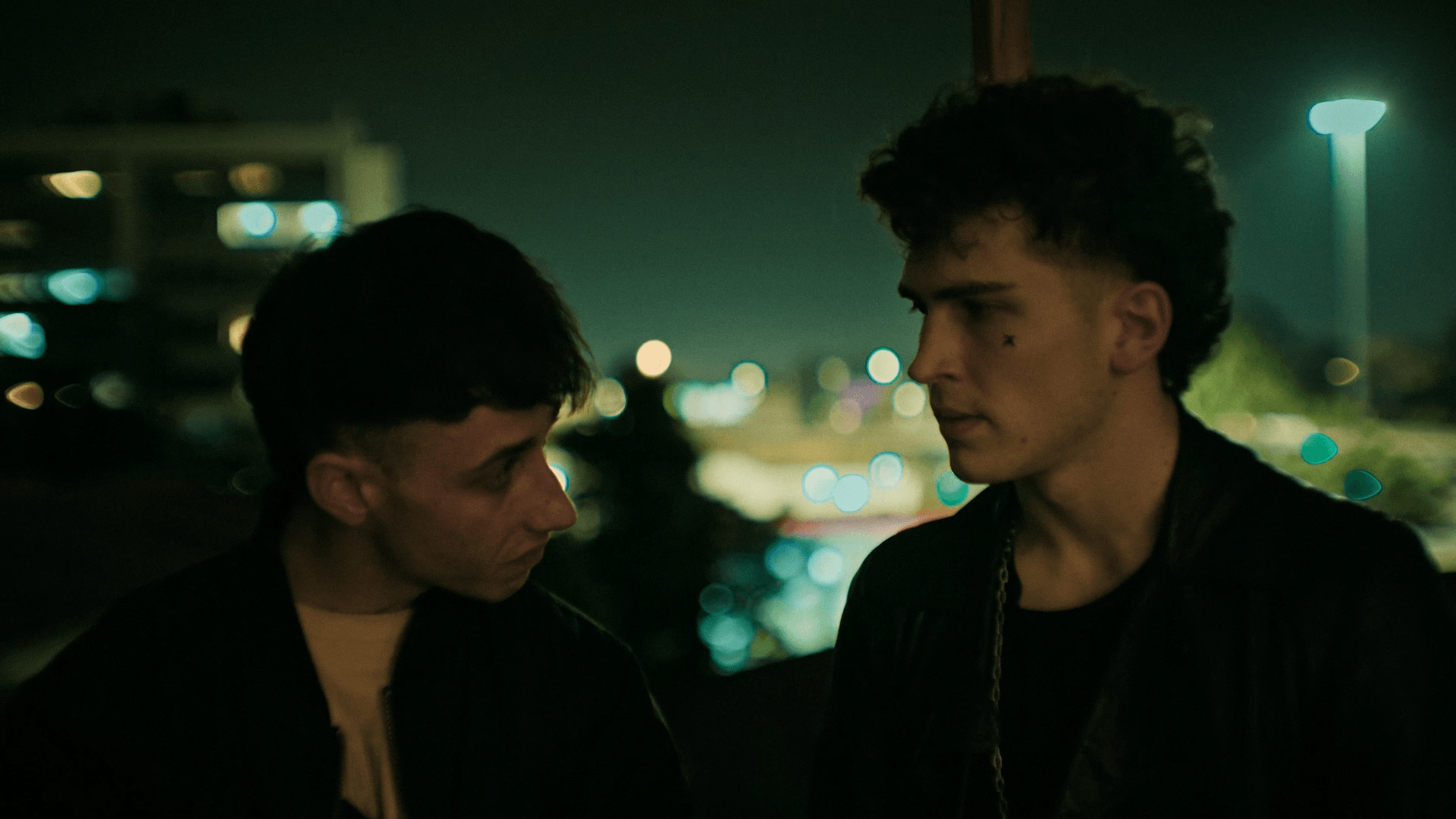 Two young men face each other in a nighttime setting with blurred city lights in the background.