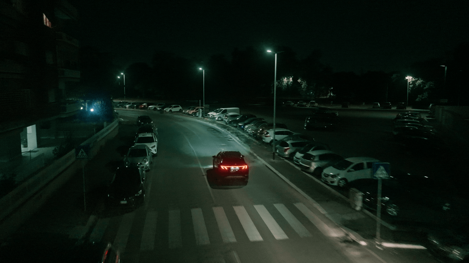 Car driving on a street with parked cars and streetlights at night, crosswalk visible, dark sky.