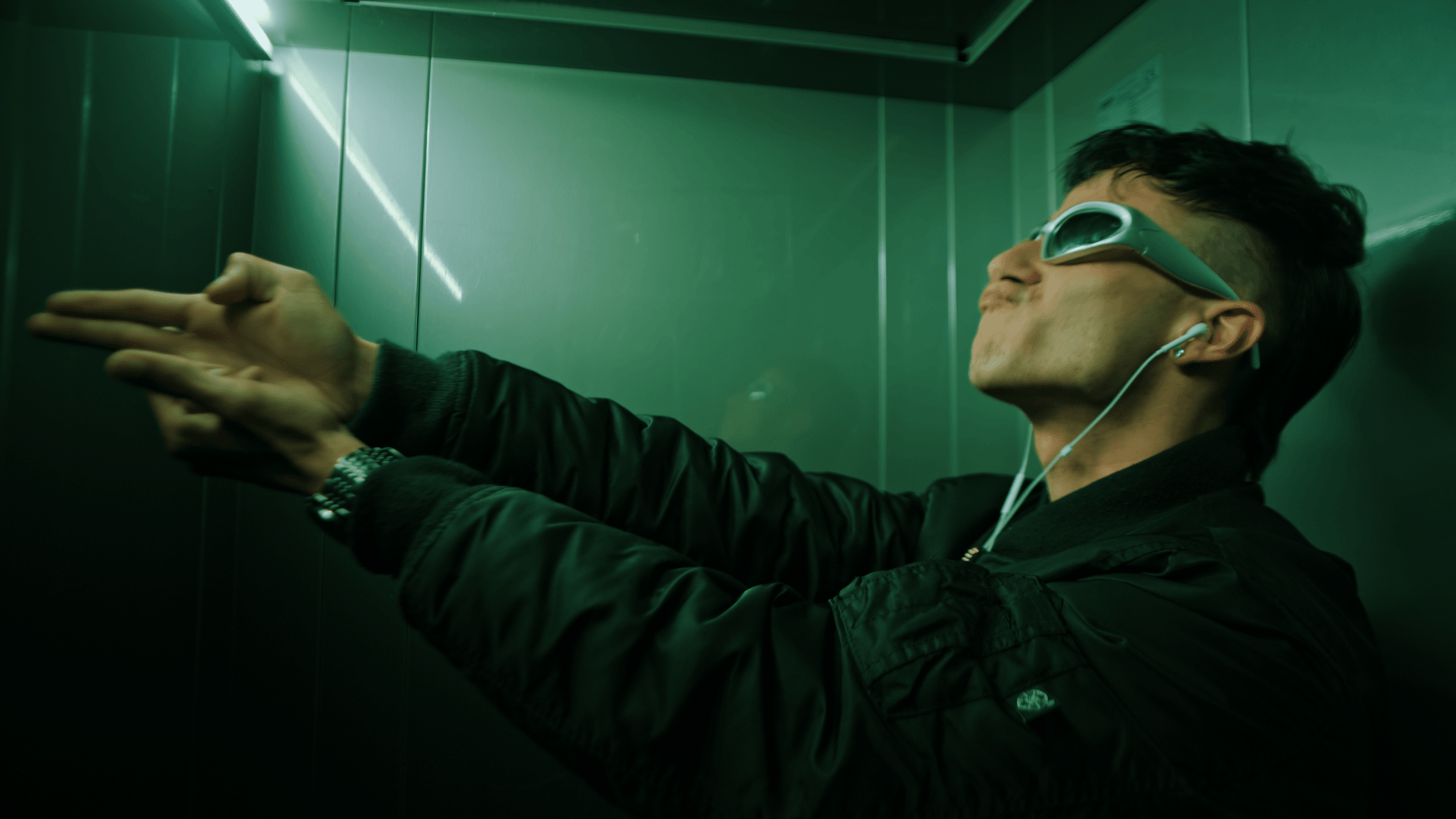 Young man in an elevator wearing VR goggles, with hands extended forward, in a dark jacket, green walls, and reflective surfaces.