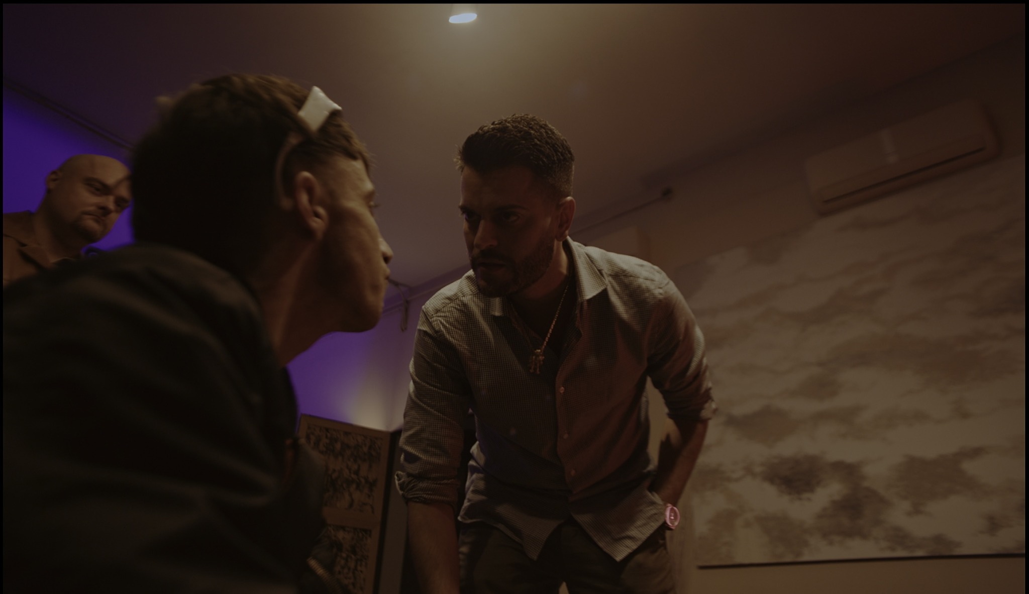Two men engaged in conversation in a dimly lit room with a patterned wall, other people in the background.