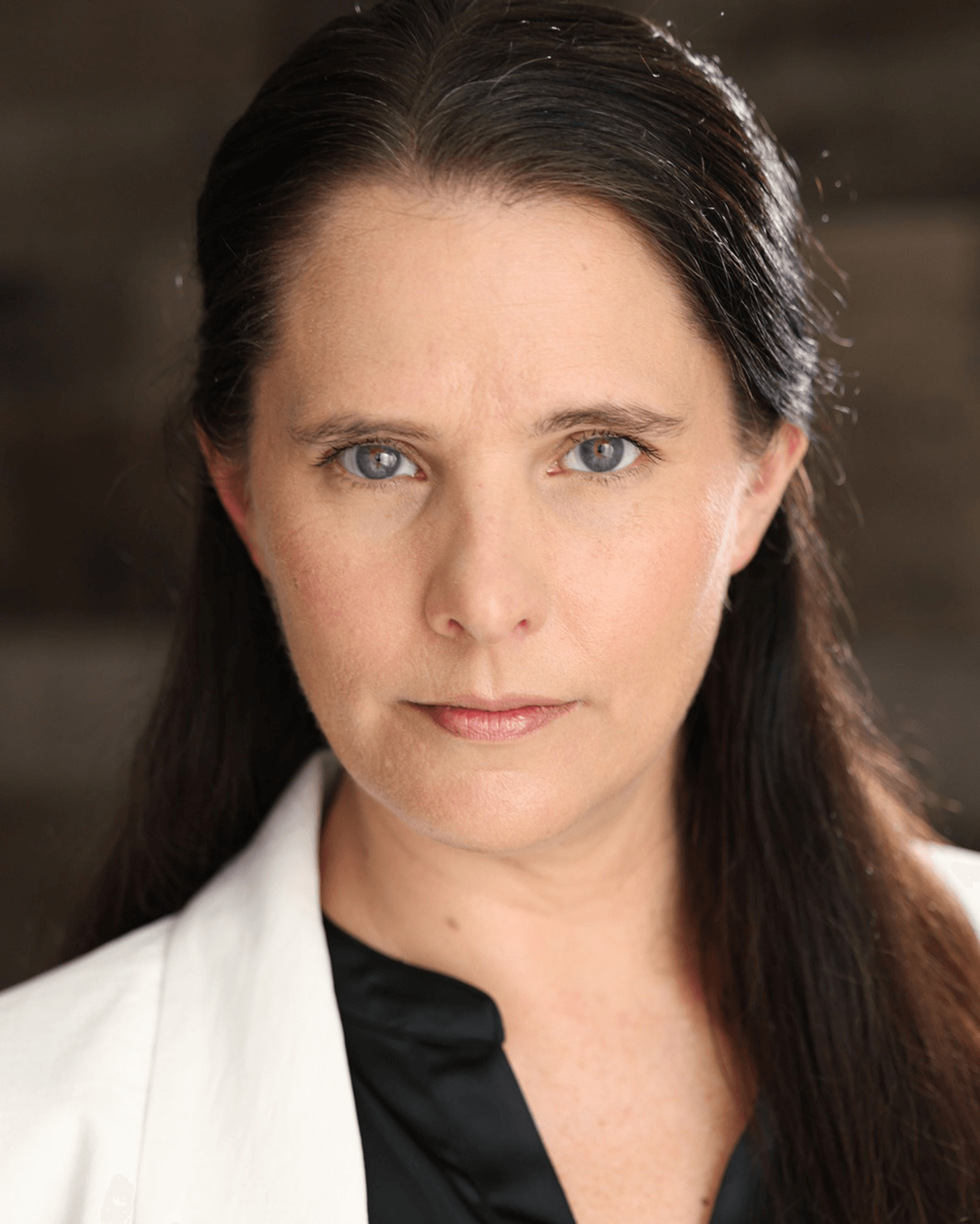 Woman with long dark hair and blue eyes looking directly at the camera, wearing a white top and black scarf, neutral background