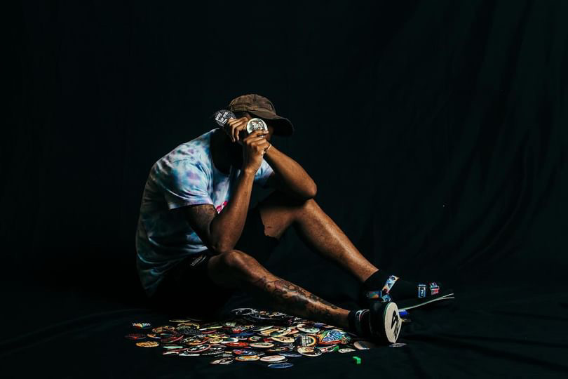 Person sitting on the floor with a black background, wearing a hat, holding a phone, surrounded by scattered items.