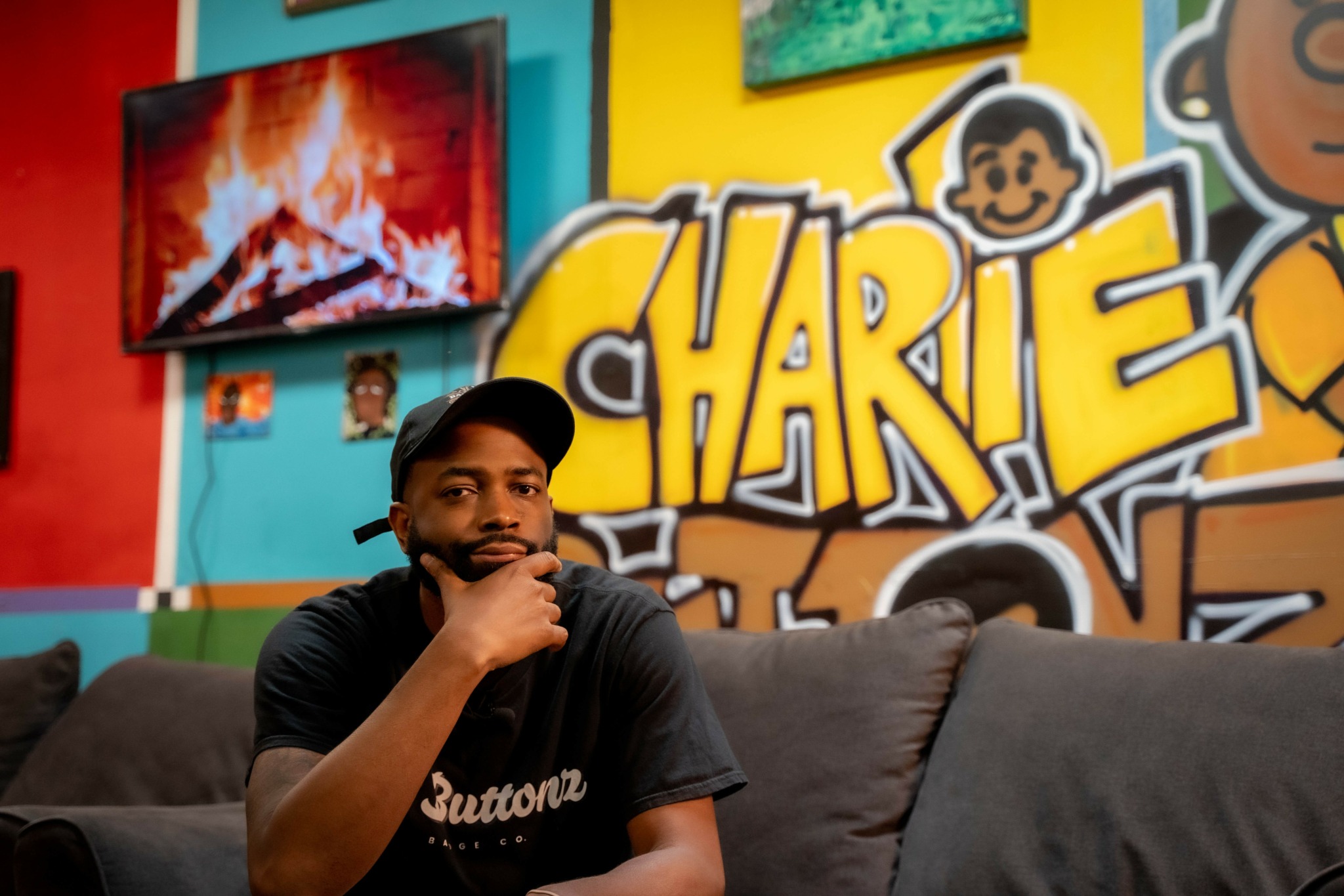 Young man with a beard and black cap sitting on a couch in front of colorful graffiti wall, including a large 'CHARIE' sign.