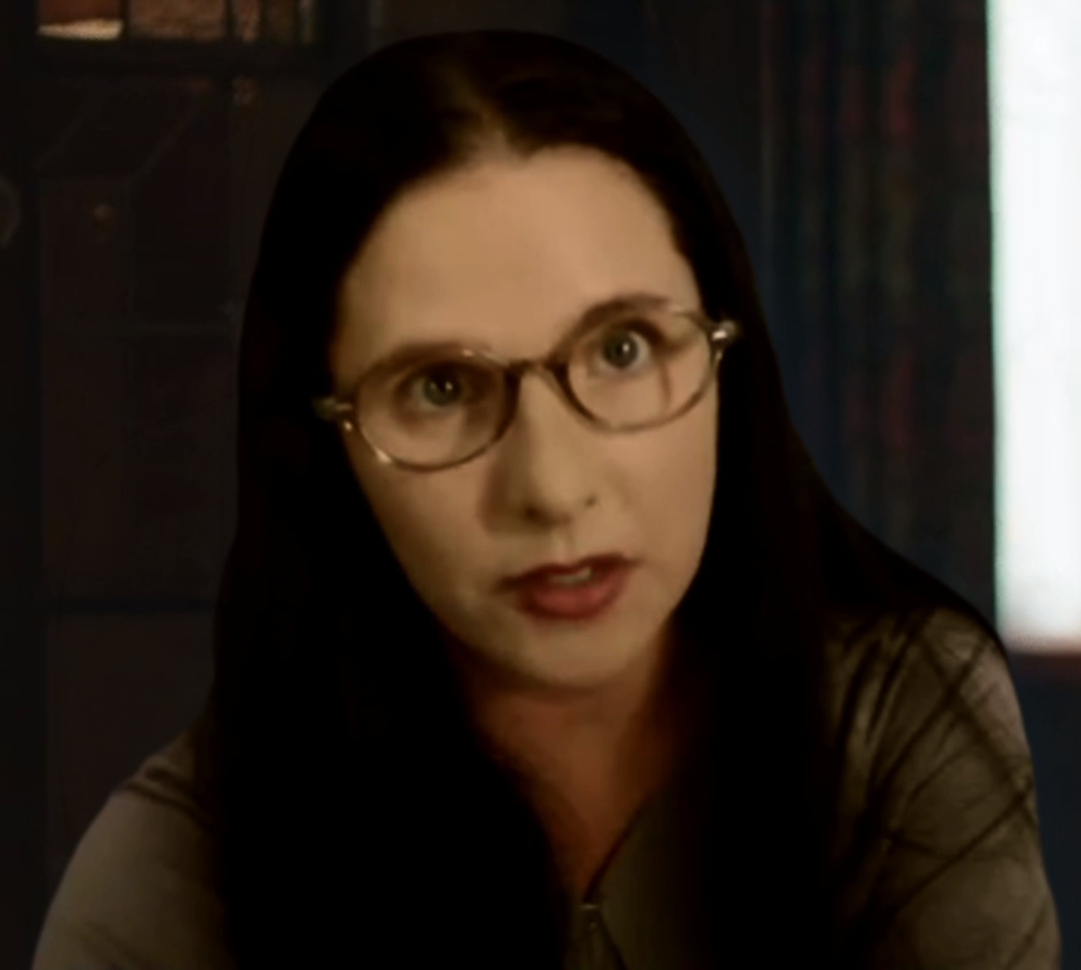 Woman with dark hair and glasses looking at the camera, indoors with dark curtains in background.