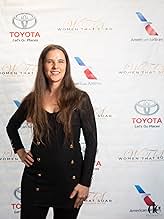 Woman with long hair smiling, standing in front of a backdrop with Toyota and American Airlines logos, wearing a black blazer.