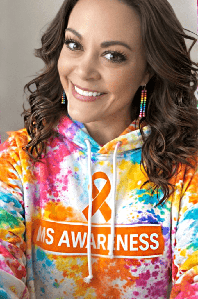 Smiling woman with curly hair wearing colorful tie-dye hoodie and rainbow earrings.