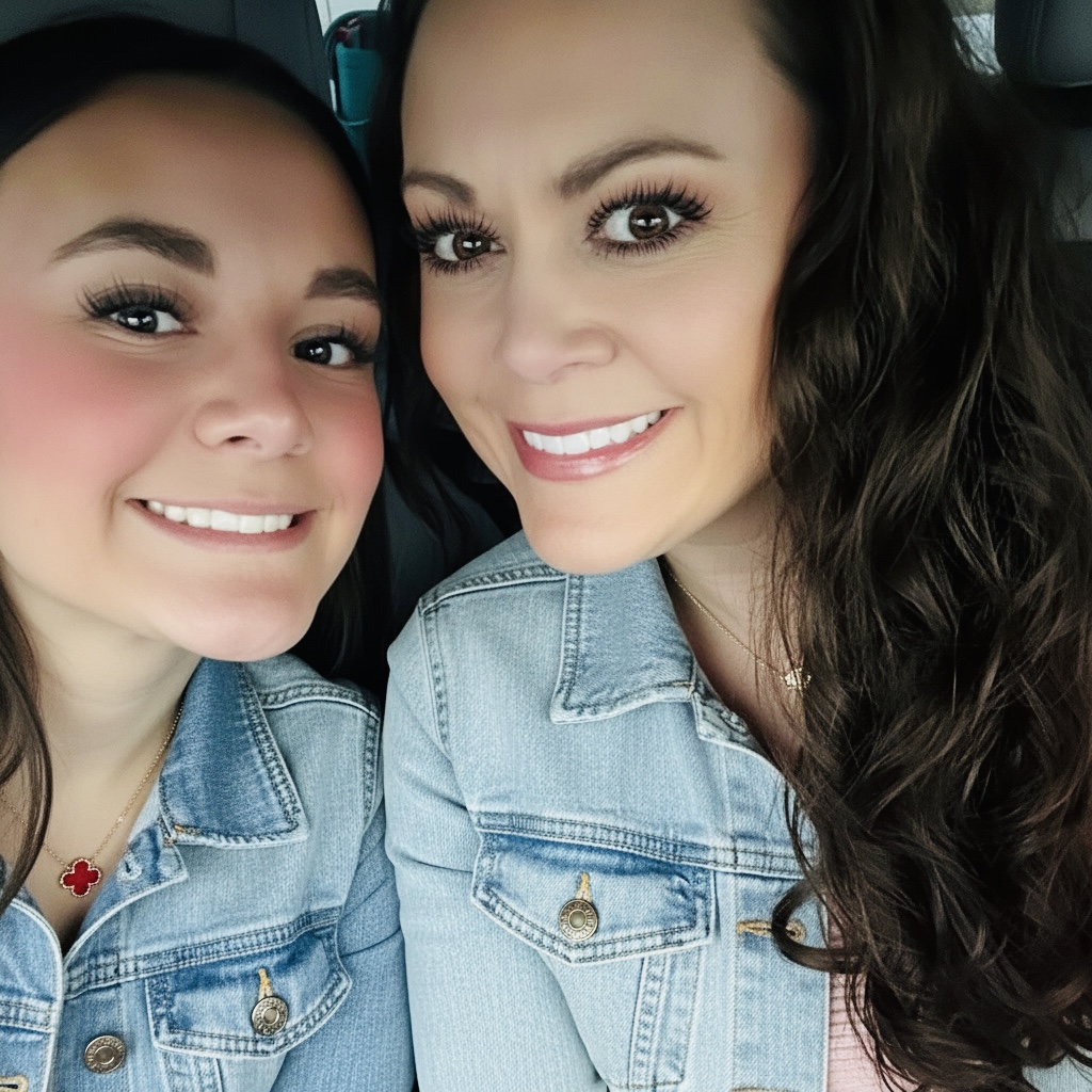 Two women smiling, wearing denim jackets, close-up selfie, dark hair, light makeup, in a vehicle.