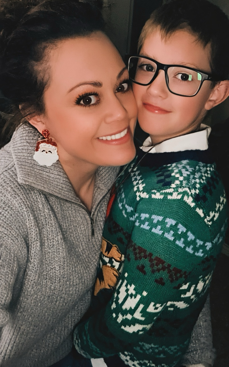 Woman and boy smiling closely, woman wearing Christmas earrings and sweater, boy with glasses and festive sweater, indoors.
