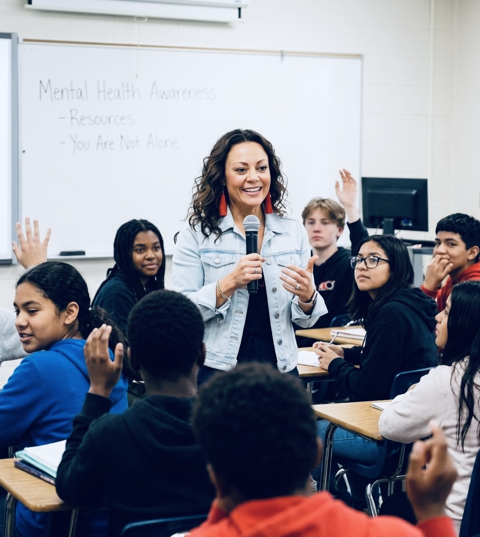 Teacher smiling and speaking to students in a classroom, some students raising hands, whiteboard in background.