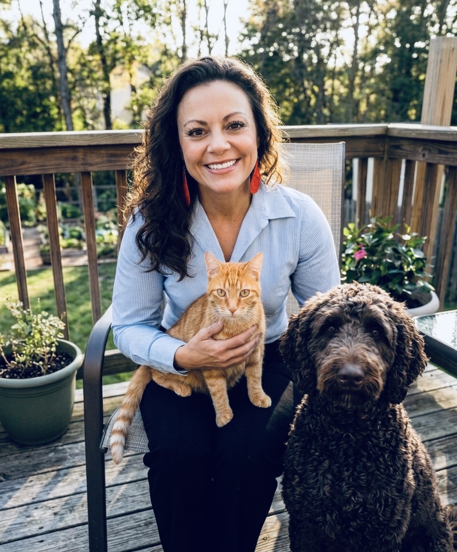 Woman sitting on a deck holding an orange cat, with a large black dog nearby, outdoors with trees and potted plants.
