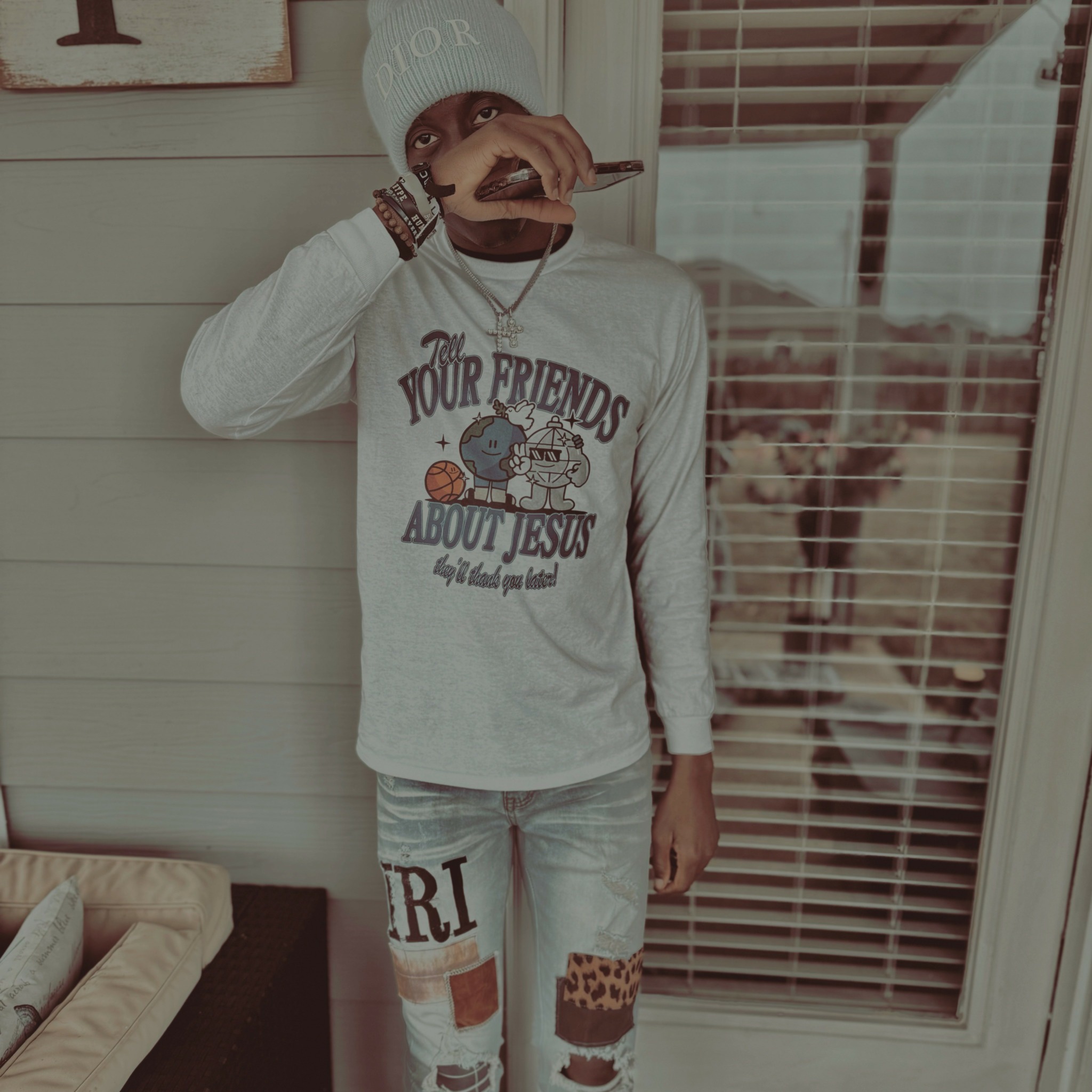Person wearing a white beanie and long-sleeve shirt, standing indoors near a window with blinds, holding a phone to their face.