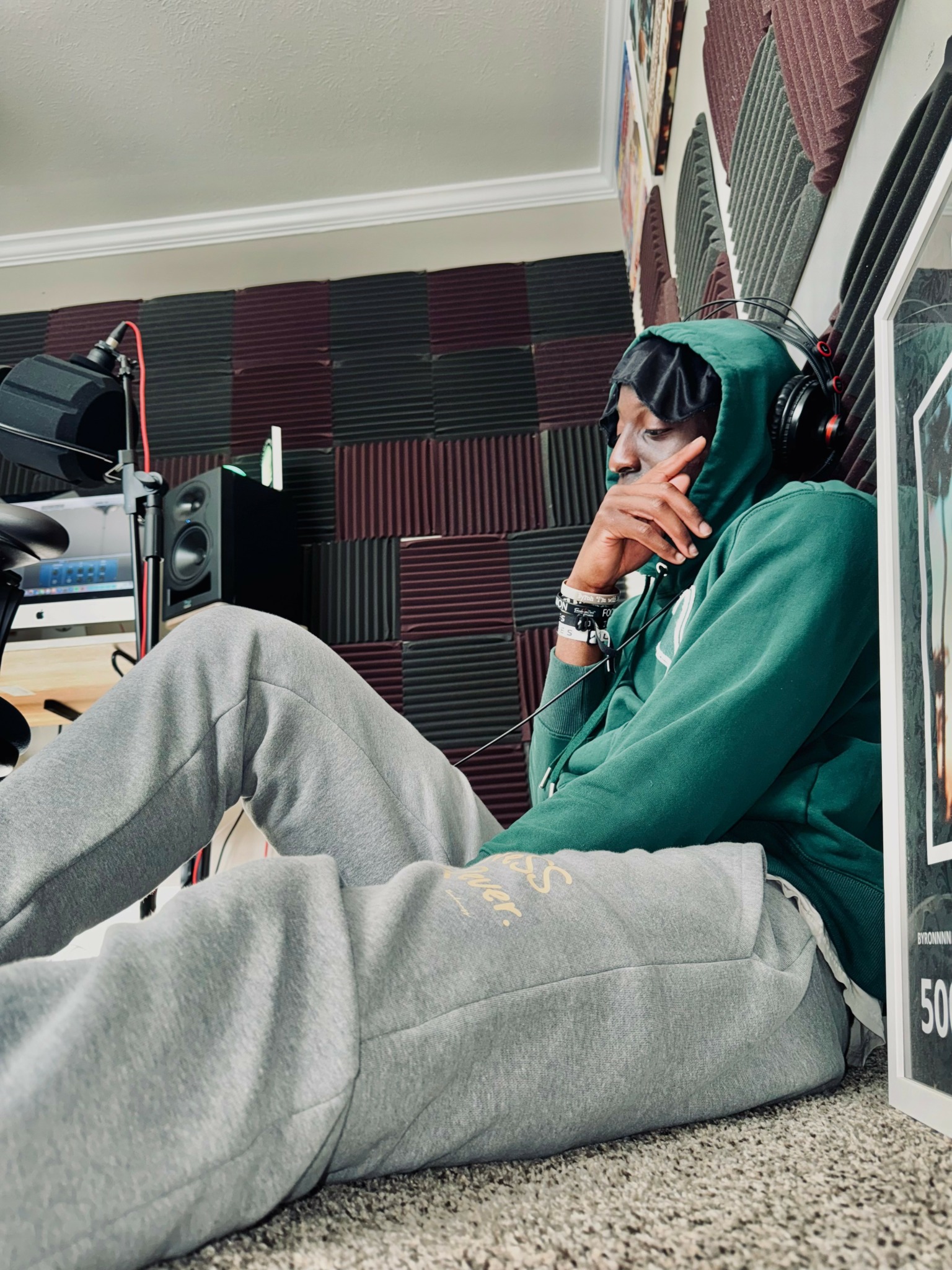 Person sitting on the floor with headphones, wearing a green hoodie and gray sweatpants, in a room with soundproofing panels.