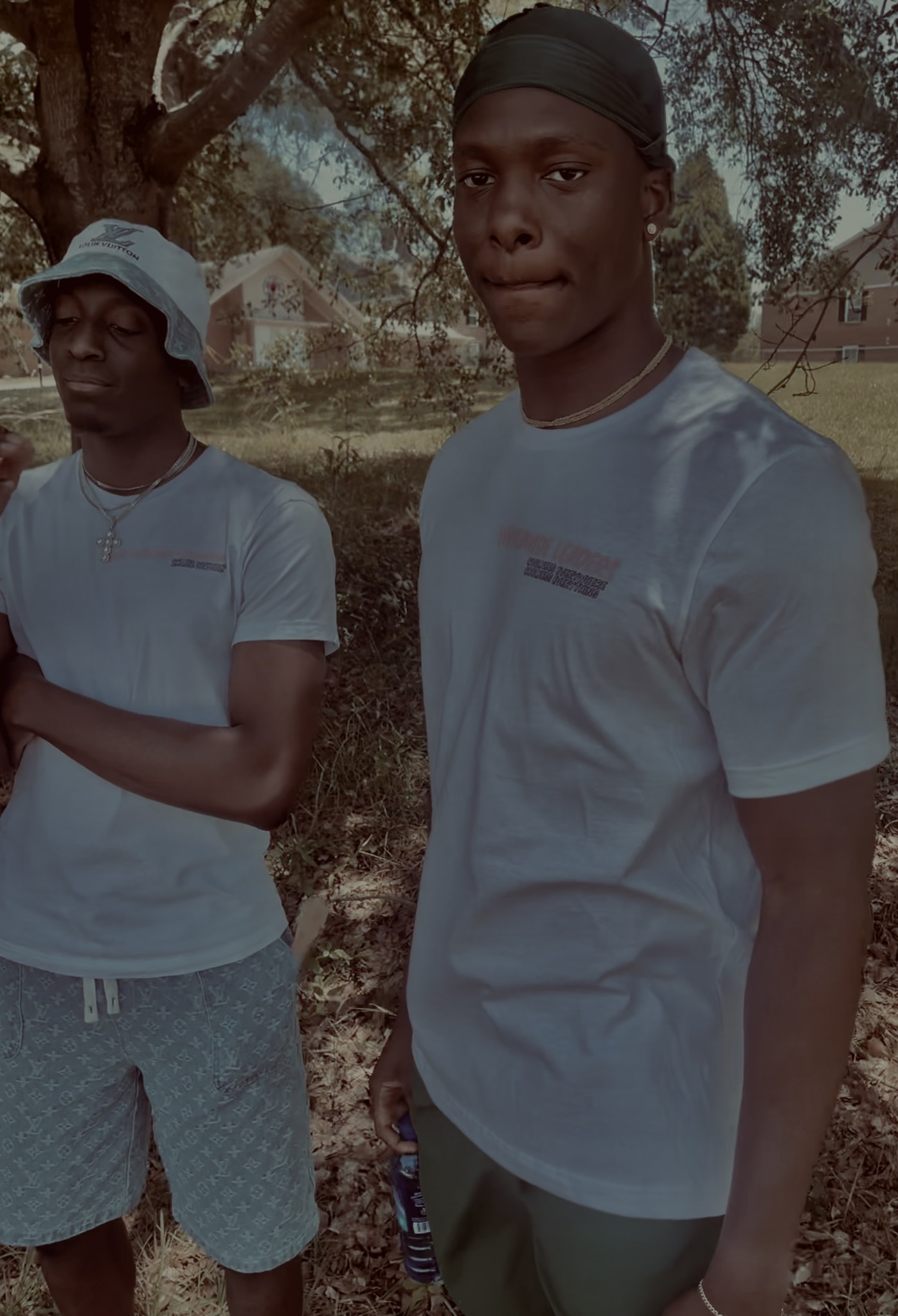 Two young men outdoors, one in a white t-shirt and the other in a light blue t-shirt, with trees in the background.