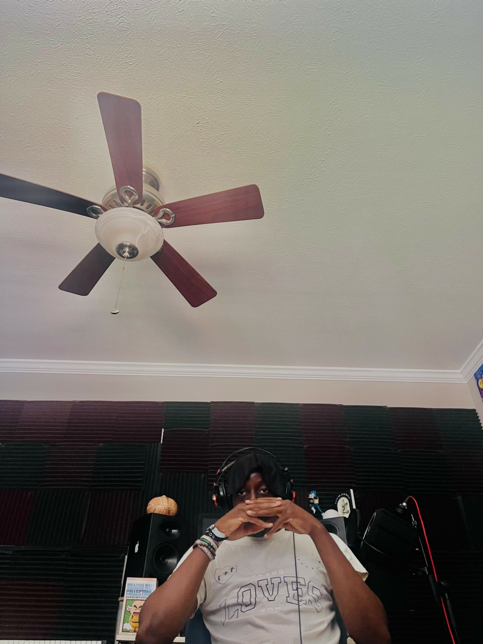 Person taking a selfie in a room with a ceiling fan and dark window blinds, holding the camera with both hands.