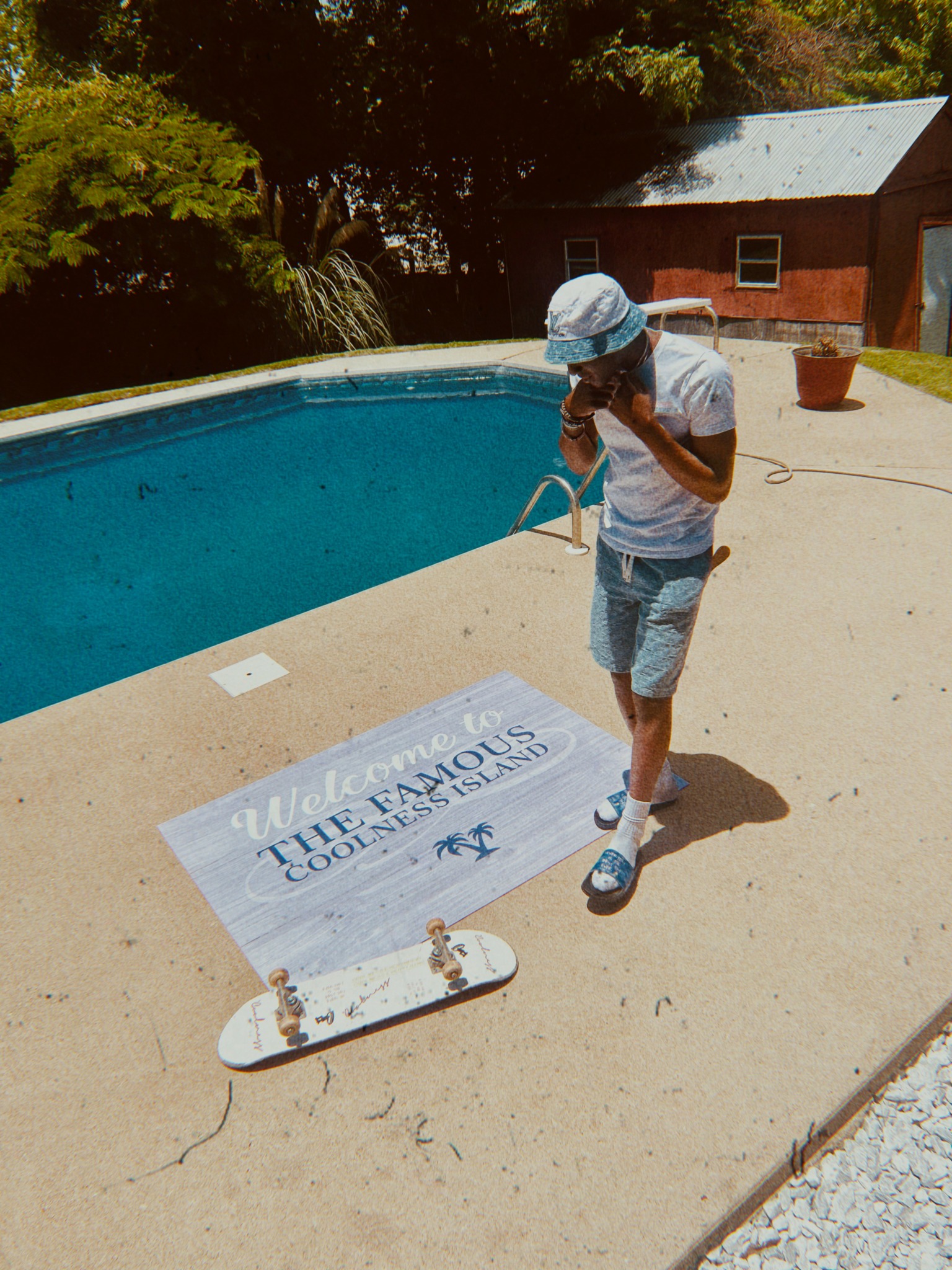 Child in a hat and shorts standing near a swimming pool, holding a towel, with a skateboard and a sign on the ground.