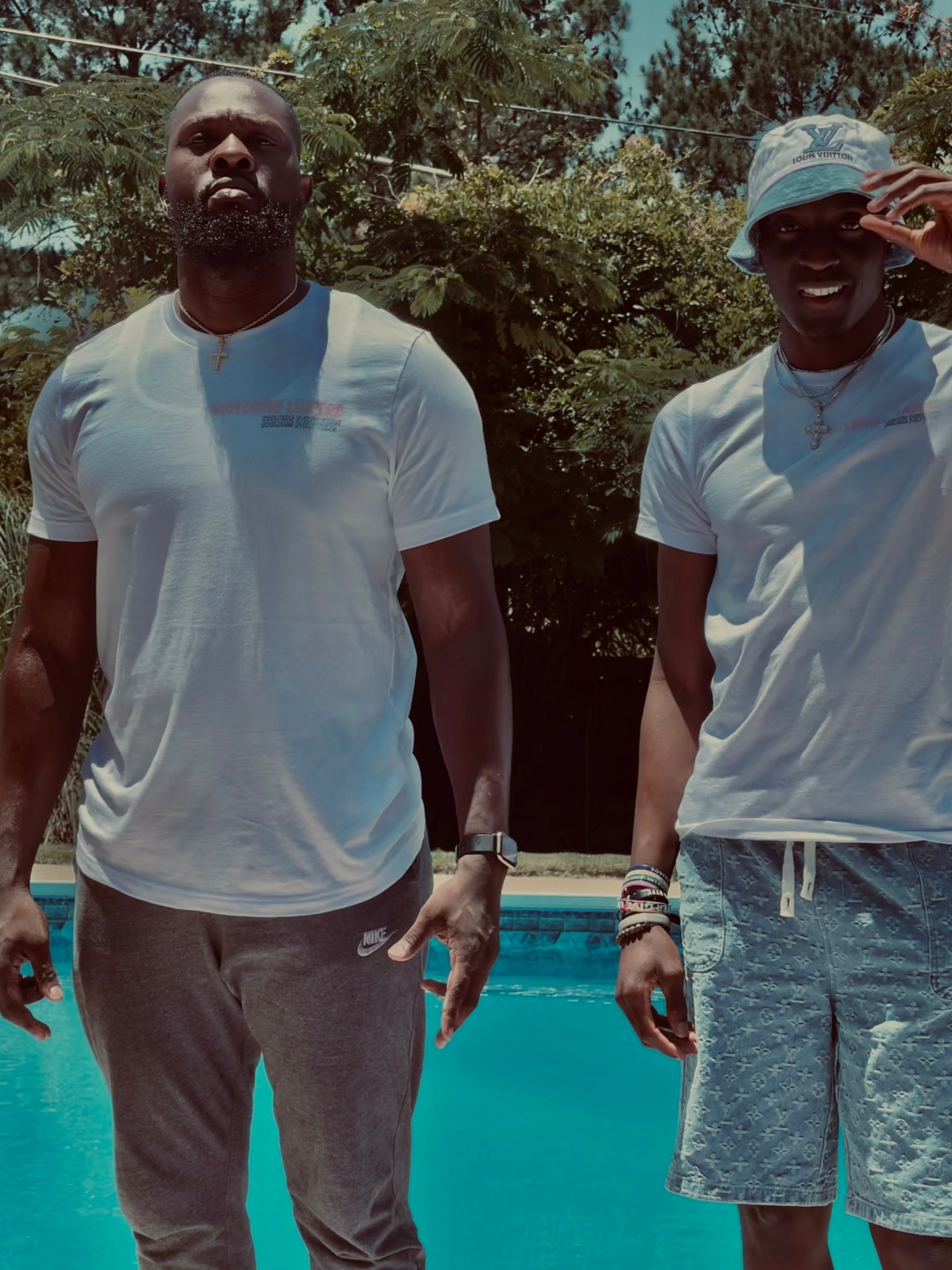 Two people standing outdoors near a pool, wearing casual clothing, with trees in the background.