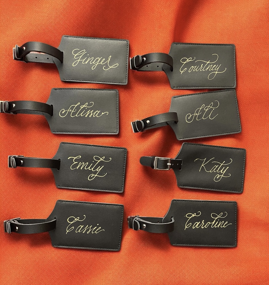 Eight black luggage tags with gold cursive names on an orange fabric background.