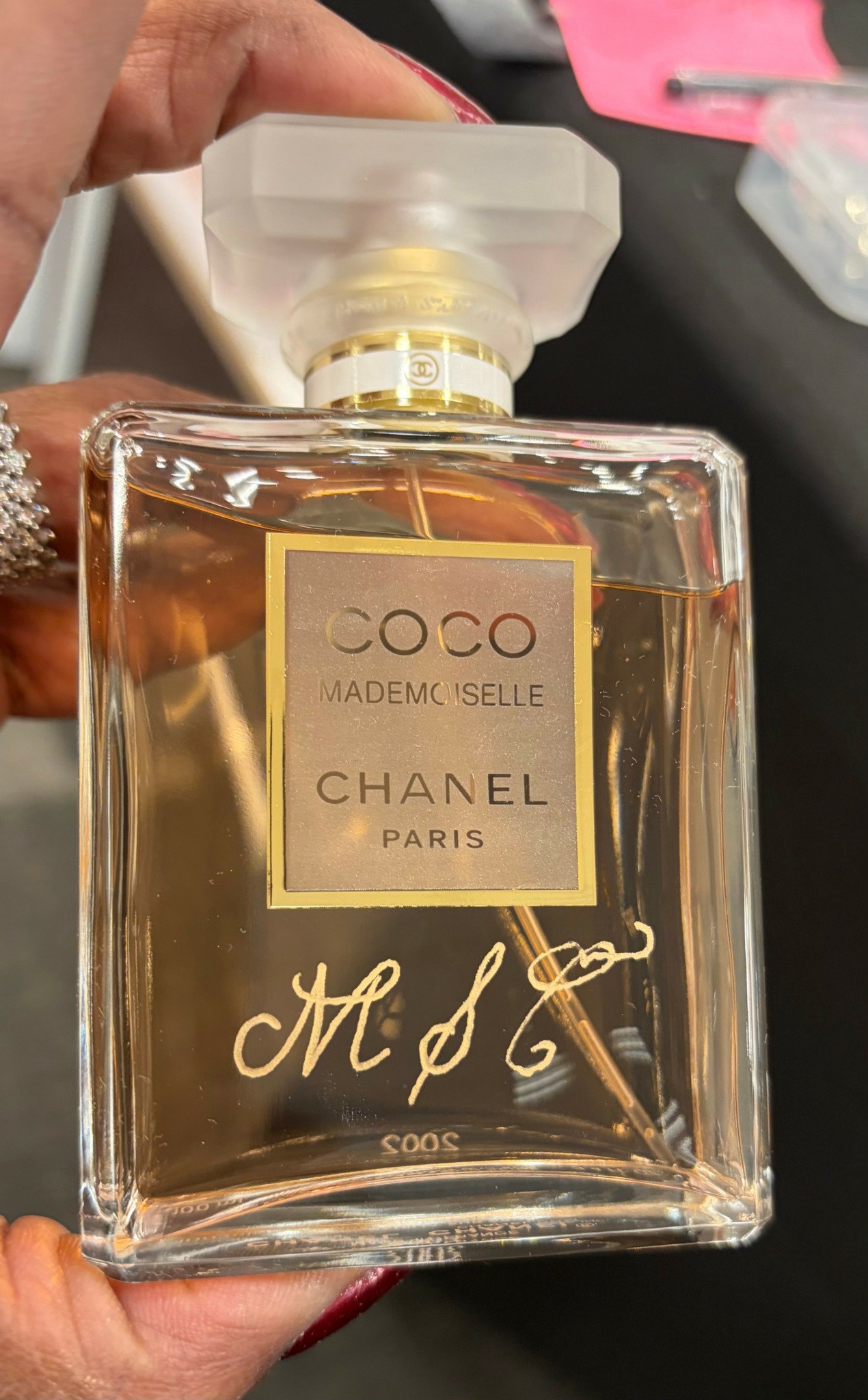 Close-up of a Chanel Coco Mademoiselle perfume bottle with a gold label, held by a hand, background blurred.