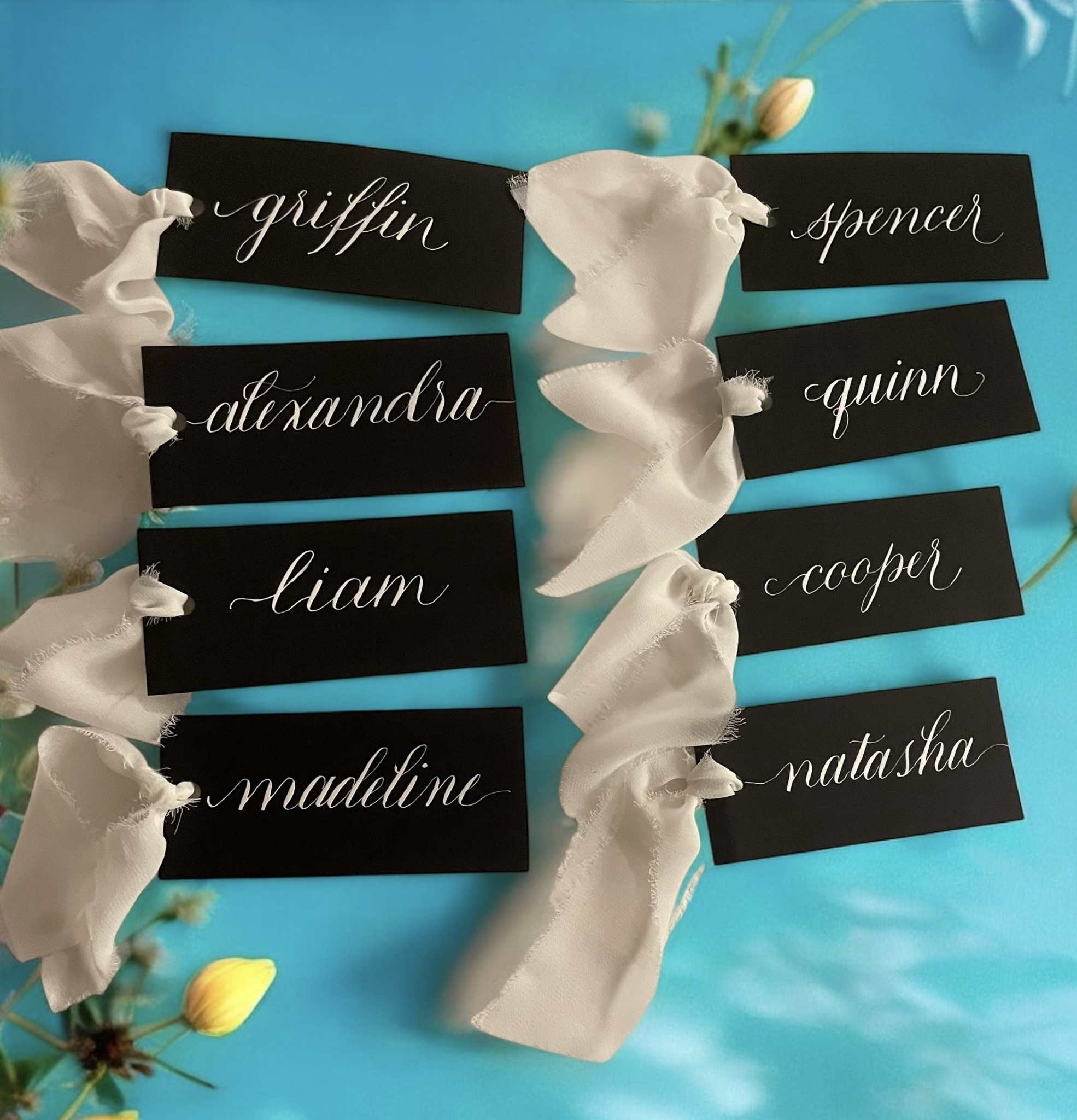 Black name tags with white cursive writing attached to white ribbons on a blue background, with some yellow flowers visible.