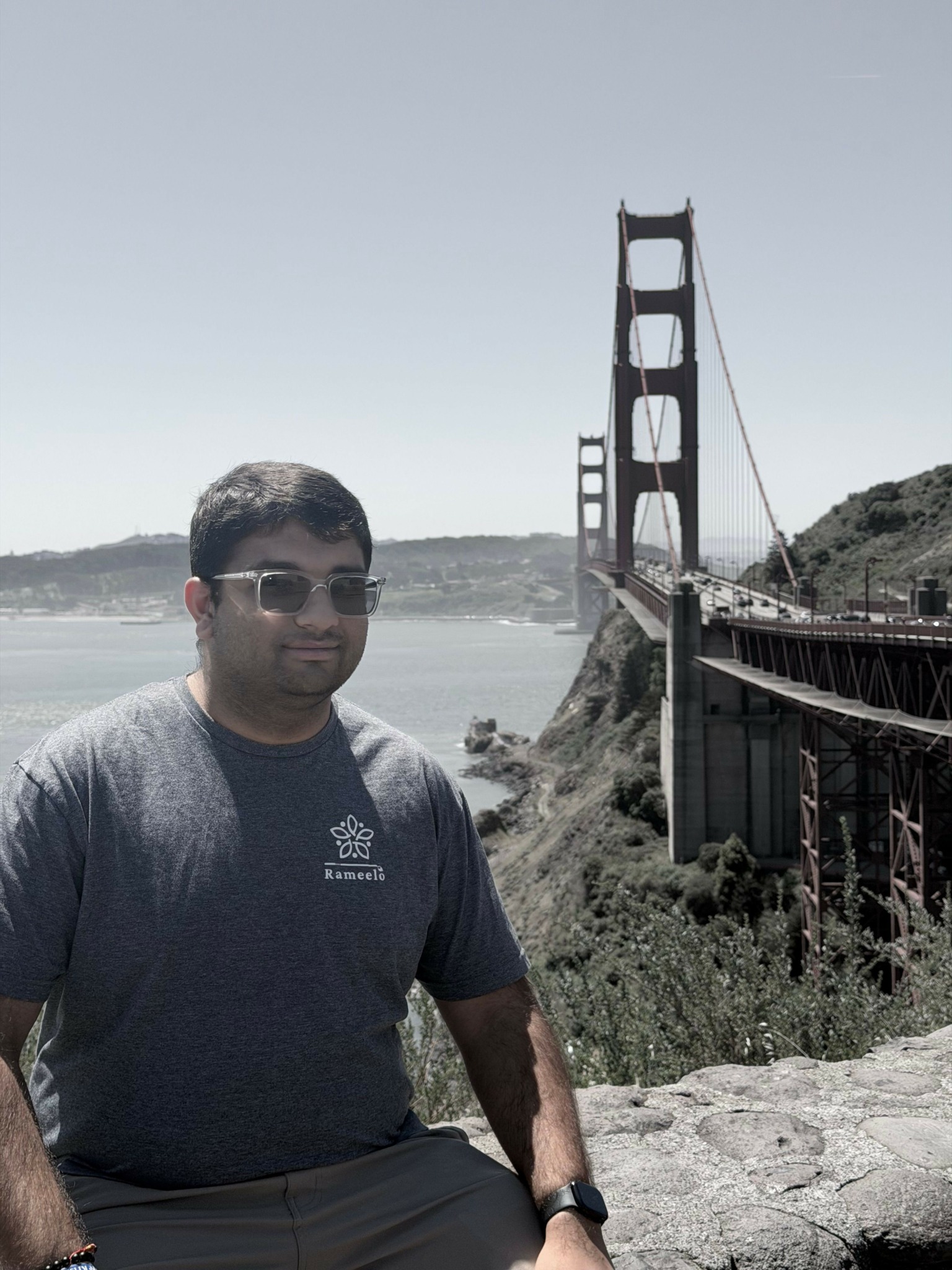 Person wearing sunglasses and a gray shirt sitting outdoors near a bridge over a body of water.
