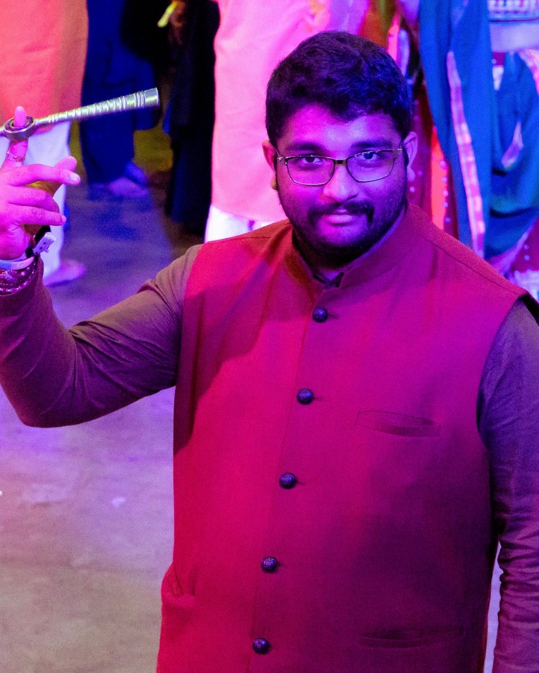 Man with glasses and a beard, wearing a maroon vest, holding a sword, at a colorful event.