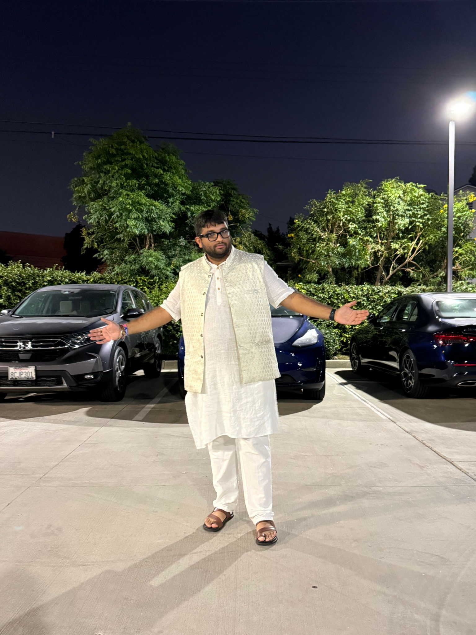 Person standing on sidewalk at night, wearing white outfit, with trees and parked cars in background.