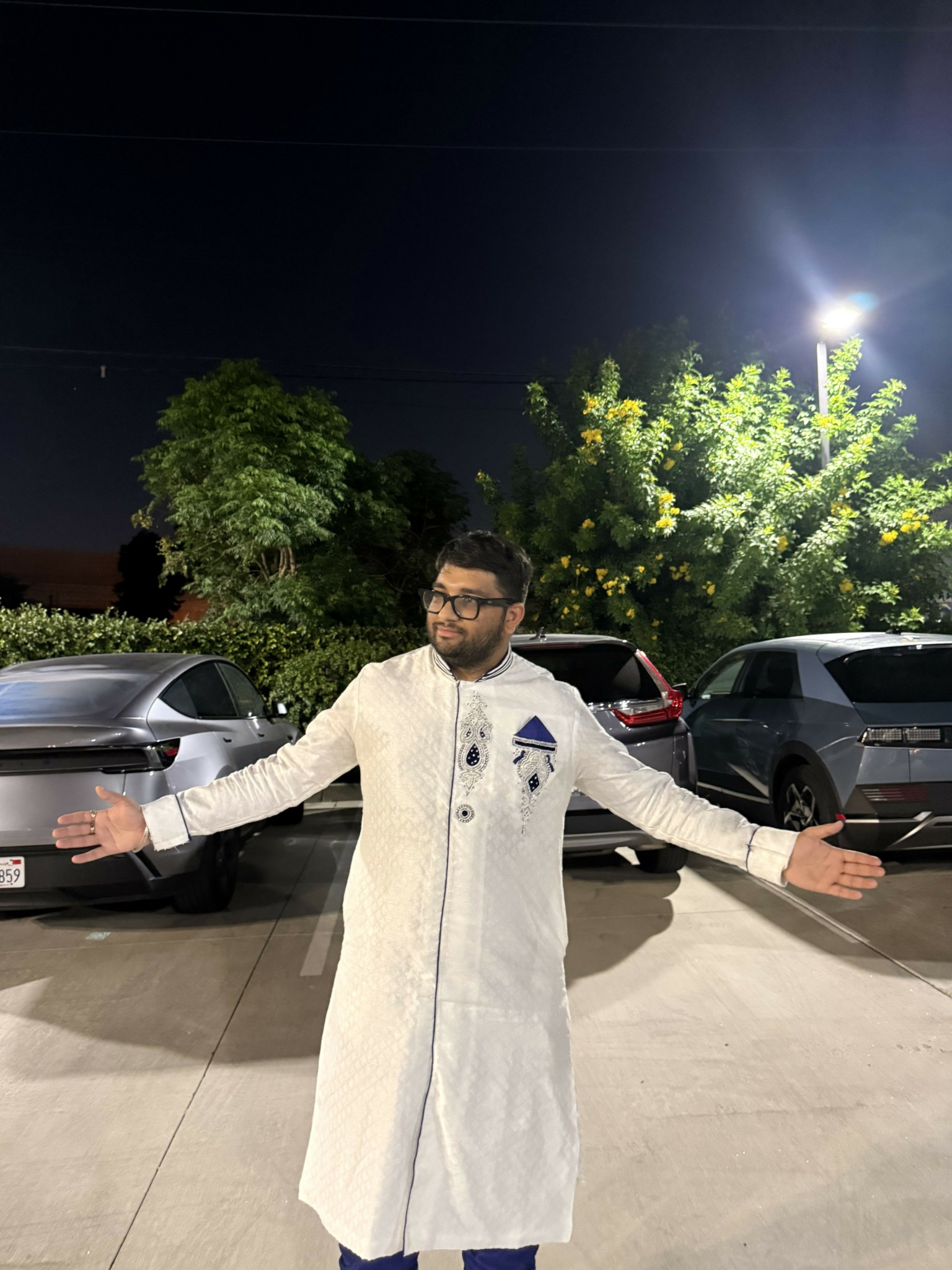 Man in traditional attire standing outdoors at night with arms outstretched, cars and trees in background.
