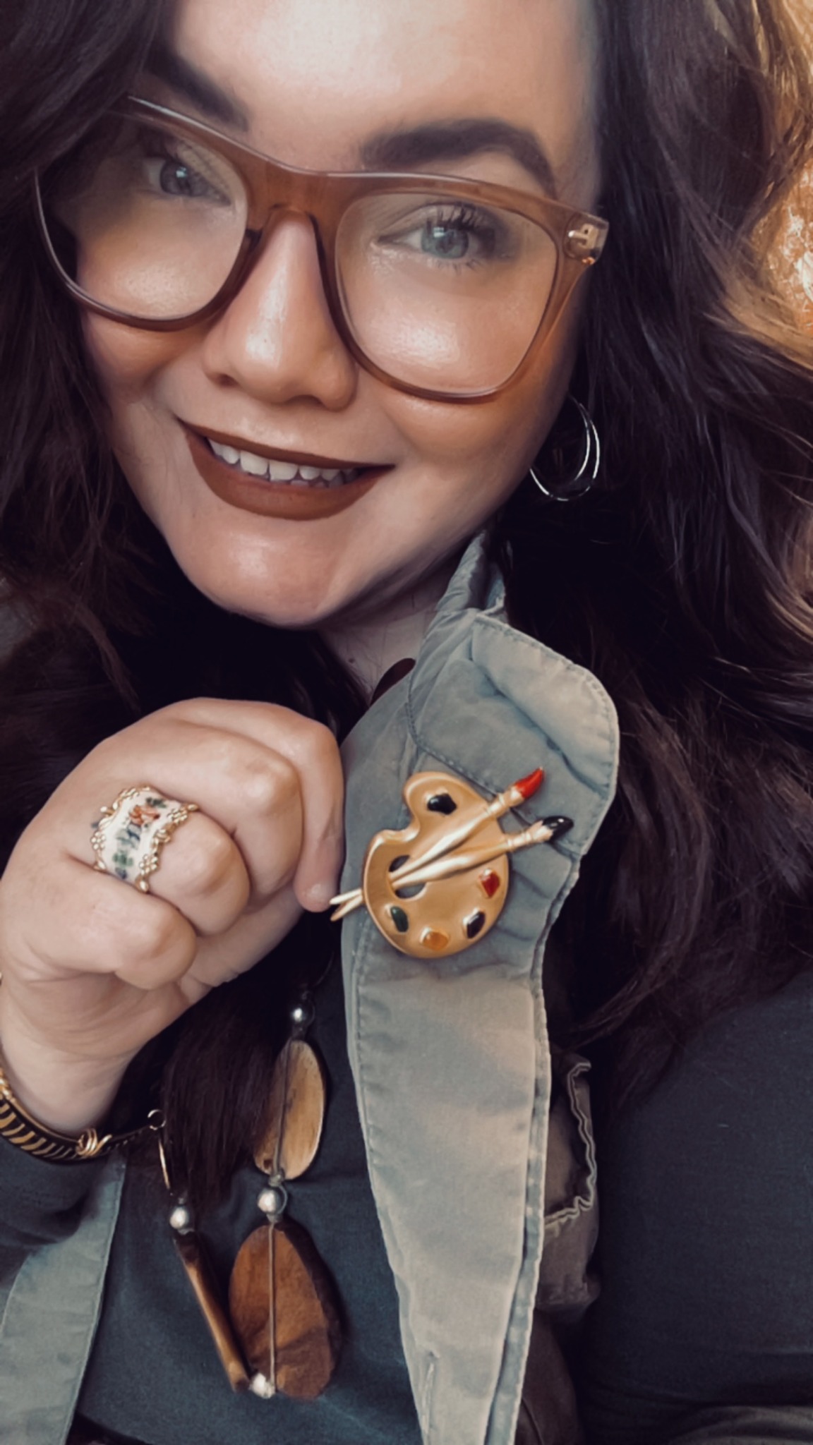 Person with glasses and dark lipstick smiling, holding a pin shaped like a guitar, wearing a green jacket.