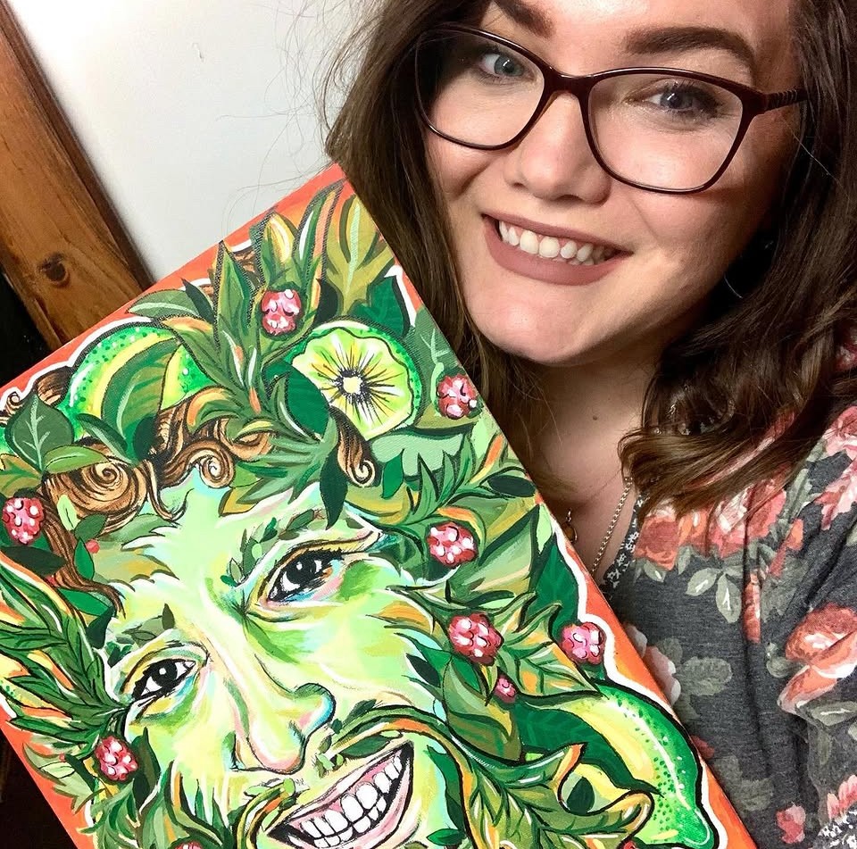 Young woman with glasses smiling, holding a colorful painting of a face surrounded by green leaves and pink flowers.