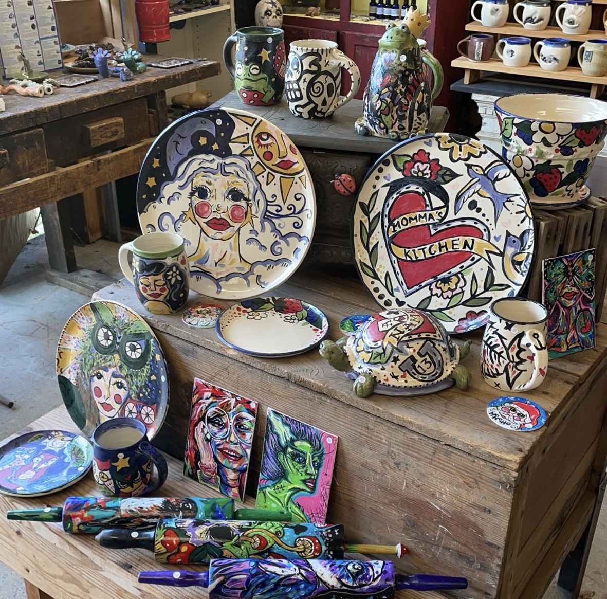 Display of colorful hand-painted ceramics, plates, mugs, and artwork on a wooden table in a shop.
