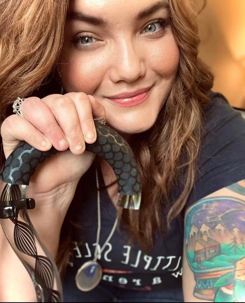 Young woman with wavy hair smiling, holding a black and gray textured object, wearing a dark shirt and colorful tattoo sleeve.