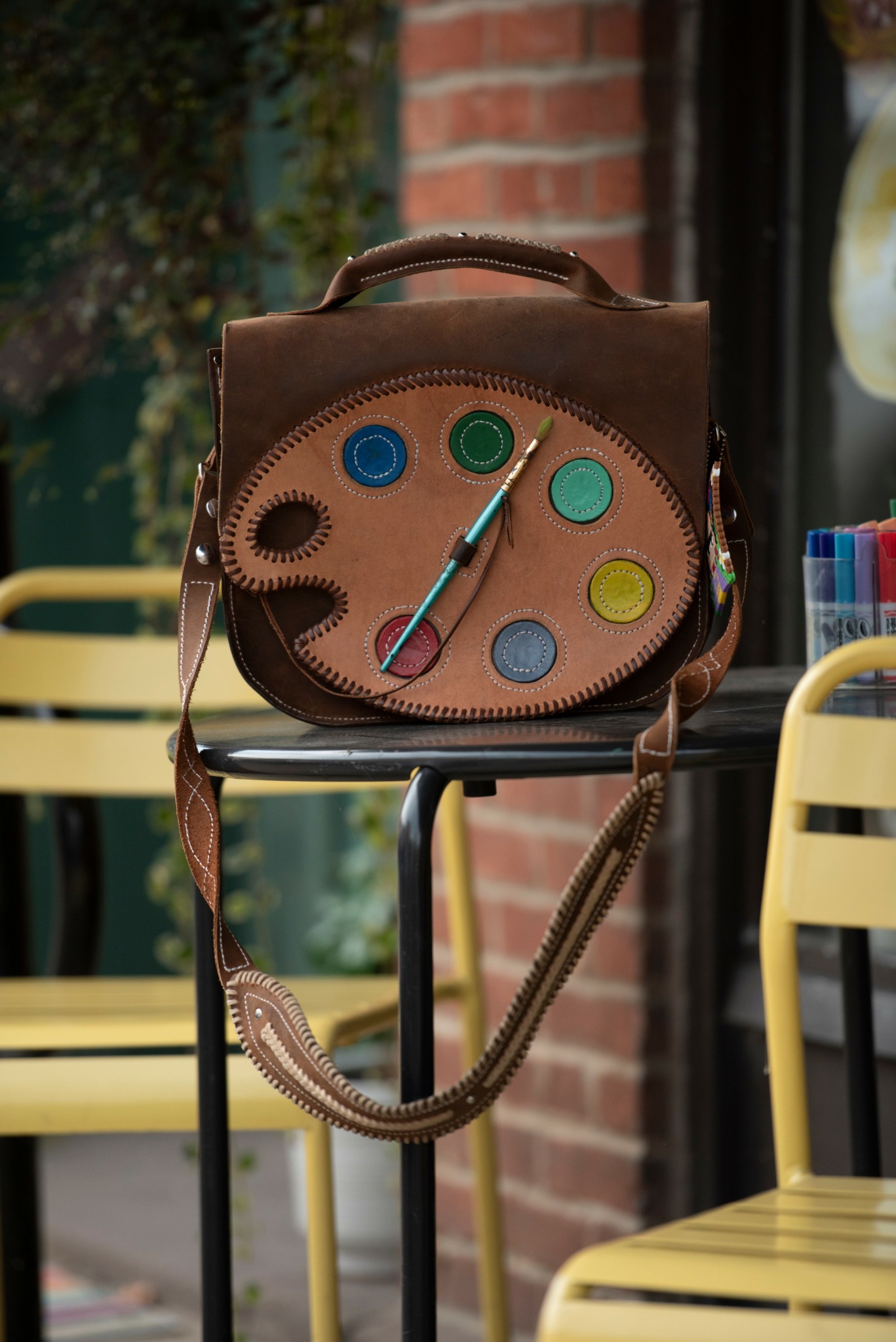Brown bag with colorful paint palette design, placed on a black stand outdoors, with yellow chairs and a brick wall in background.