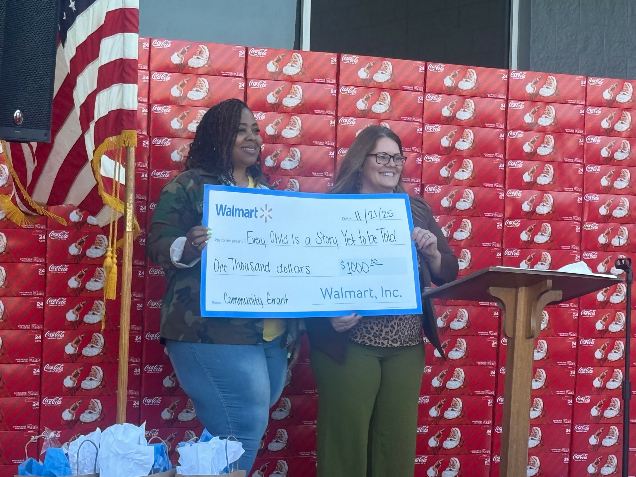 Two women holding a large check, standing in front of a red backdrop, with an American flag partially visible on the left.