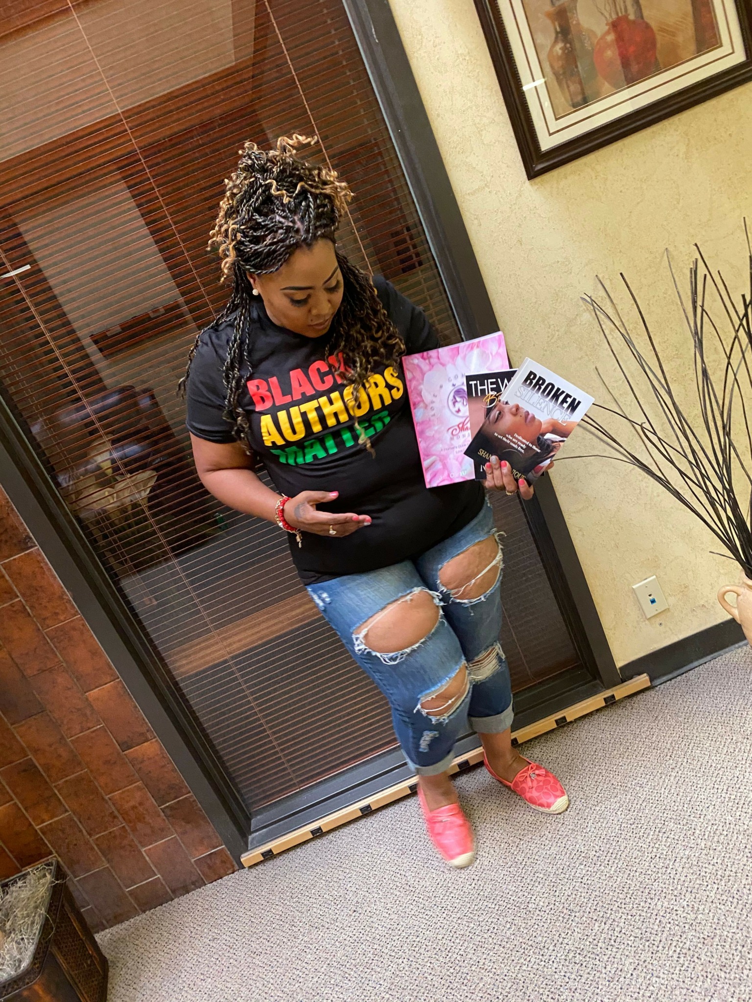 Person standing in front of glass door holding a magazine, wearing ripped jeans, black t-shirt, pink shoes, and a headscarf.