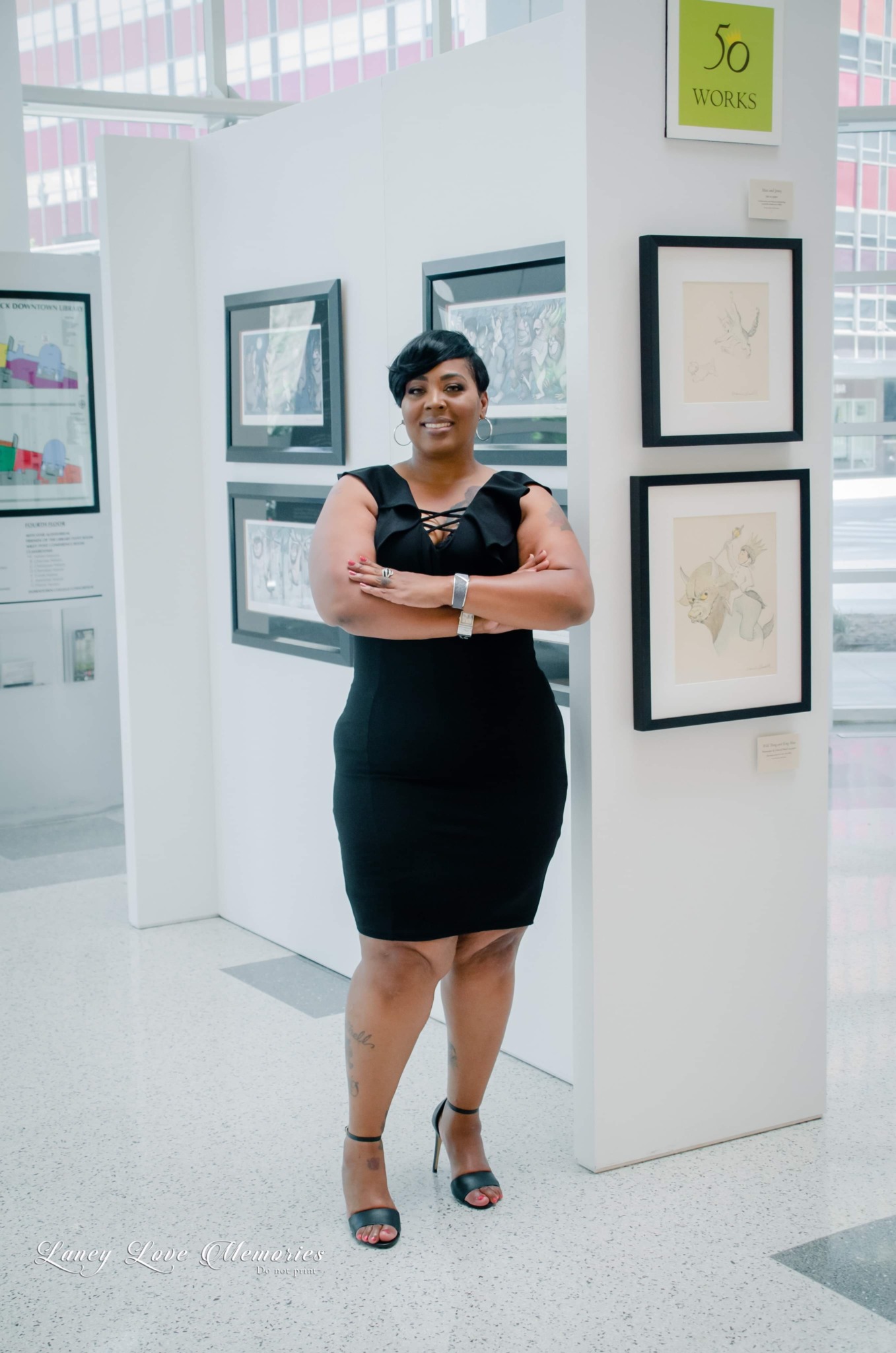 Woman in black dress standing with arms crossed in art gallery, framed artwork on white walls behind her.