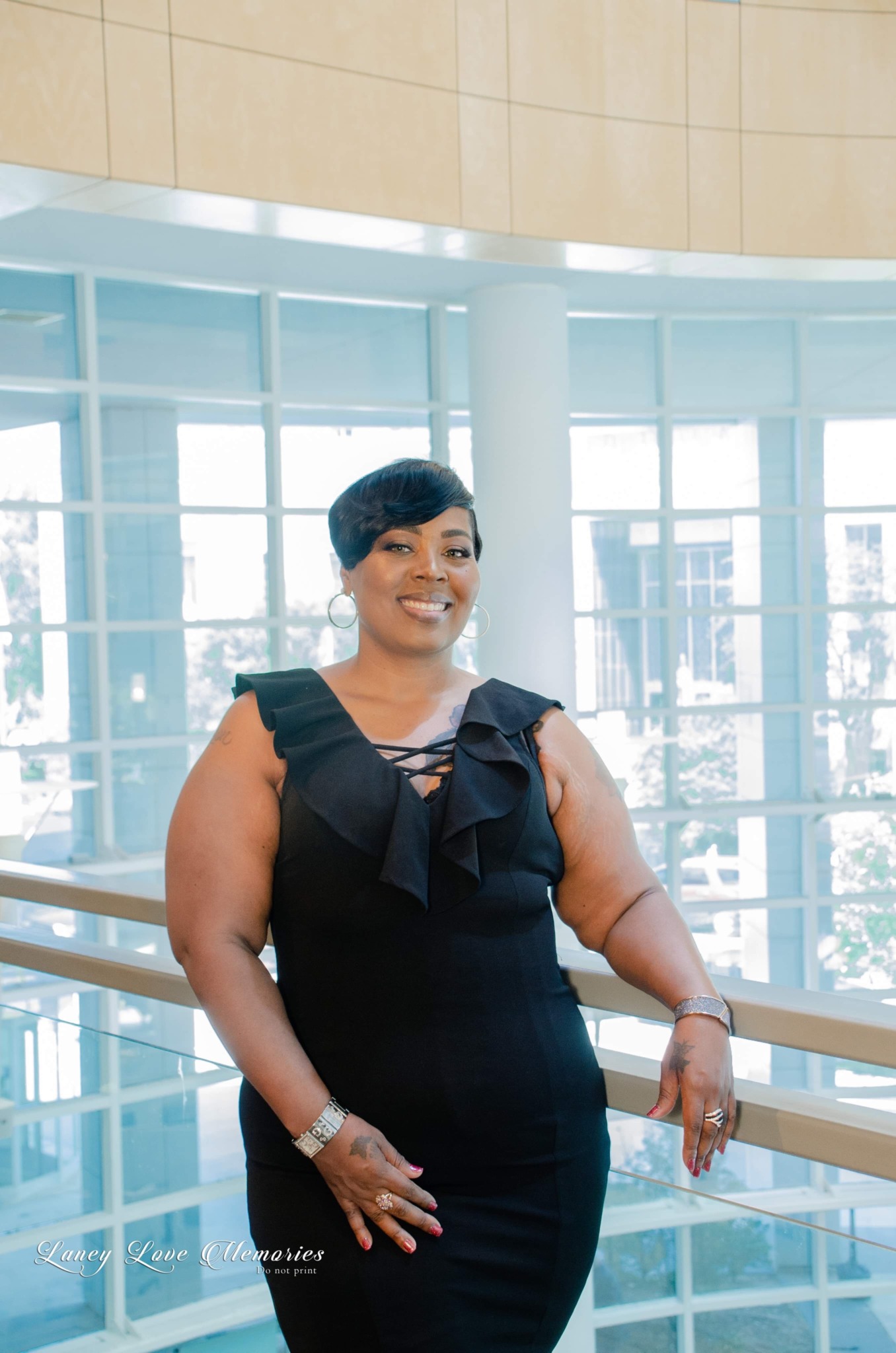 Smiling woman in a black dress with ruffled shoulders standing indoors near a glass railing, with large windows behind her.