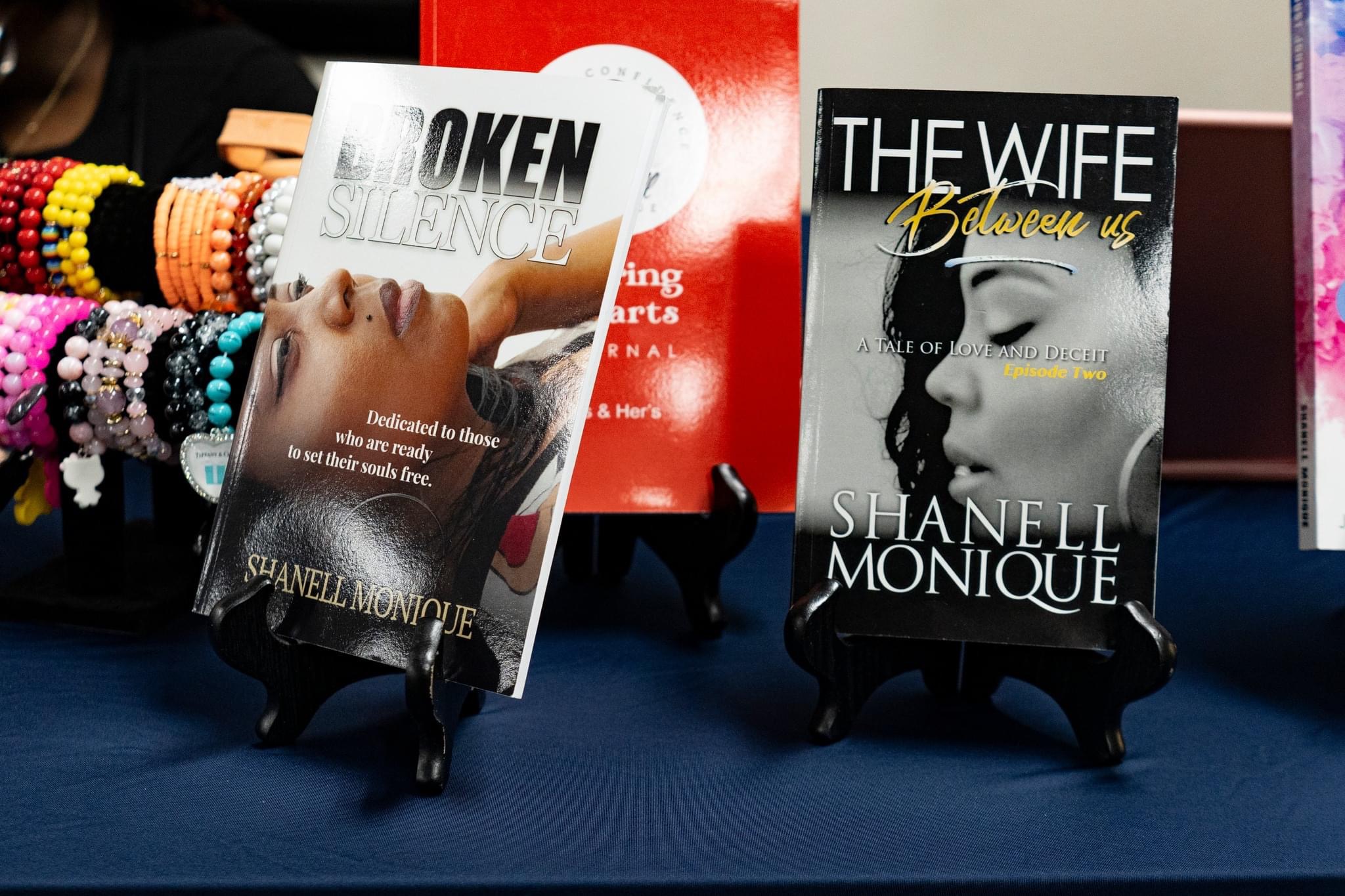 Two books displayed on black stands, one titled 'Token' and the other 'The Wife,' with colorful bracelets on the left.