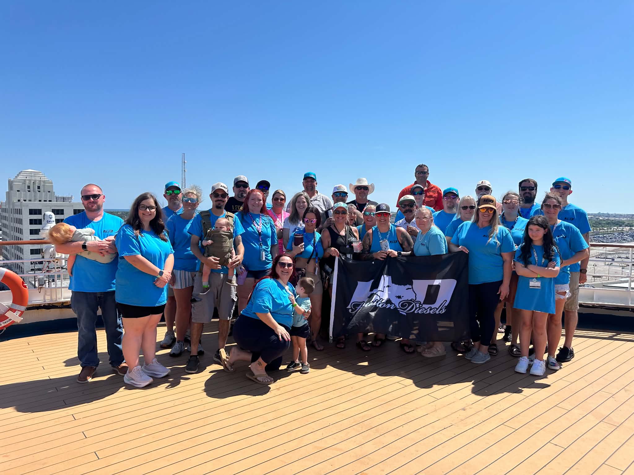 Group of people on a boat deck with city buildings in the background, some holding a banner, under a clear blue sky.